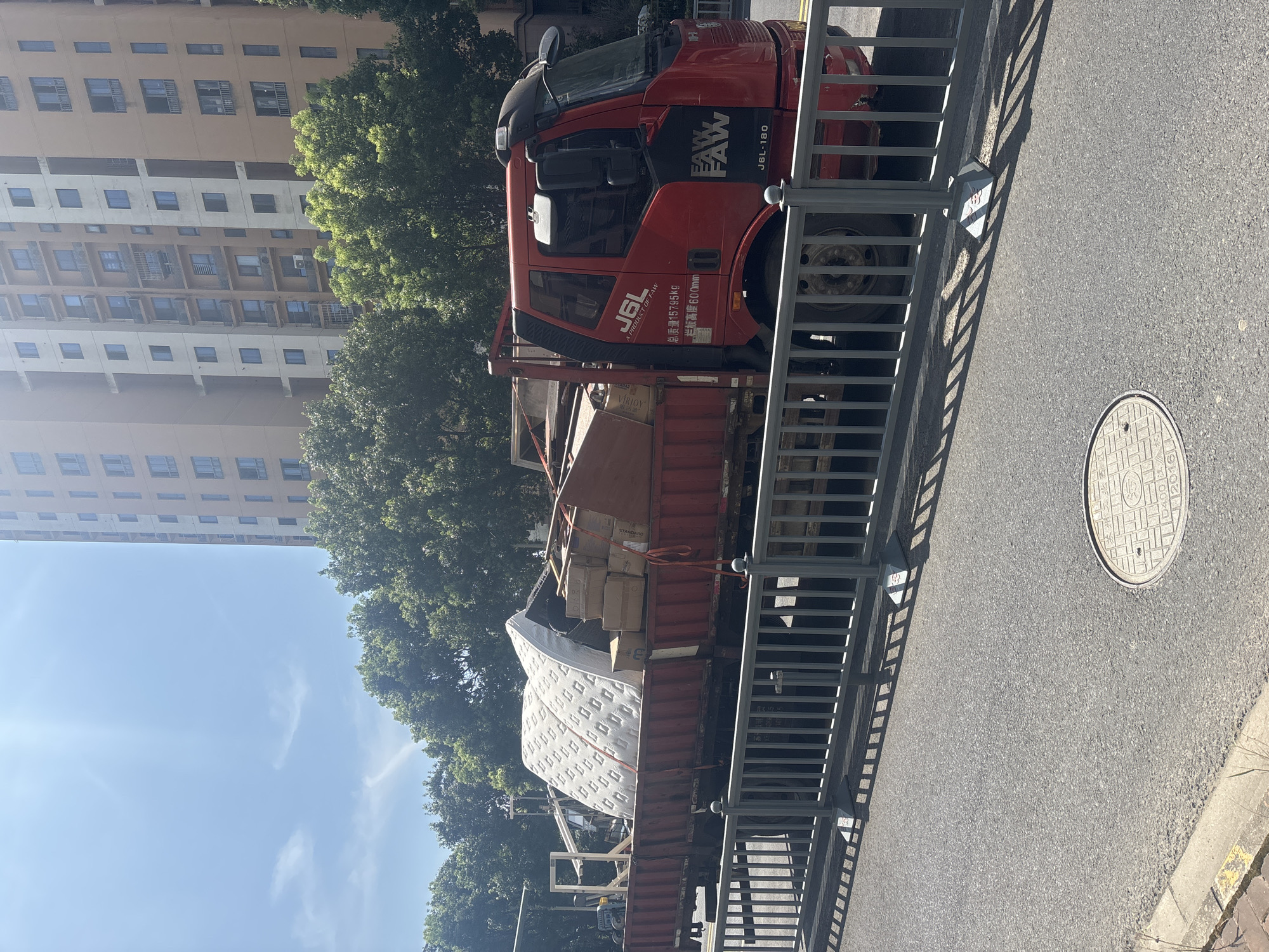 A red truck, identified by the logos "J6L" and "FAW", is parked on an asphalt road in Suzhou Industrial Park, China. The truck is loaded with various items, including a large mattress, cardboard boxes, and wooden planks. A grey metal fence runs along the side of the road in front of the truck. In the background, a tall, multi-story building with many windows stands amidst lush green trees. The sky is bright blue with a few wispy clouds. The sun is shining, casting shadows from the fence onto the road. A circular manhole cover is visible on the road near the bottom right of the image.