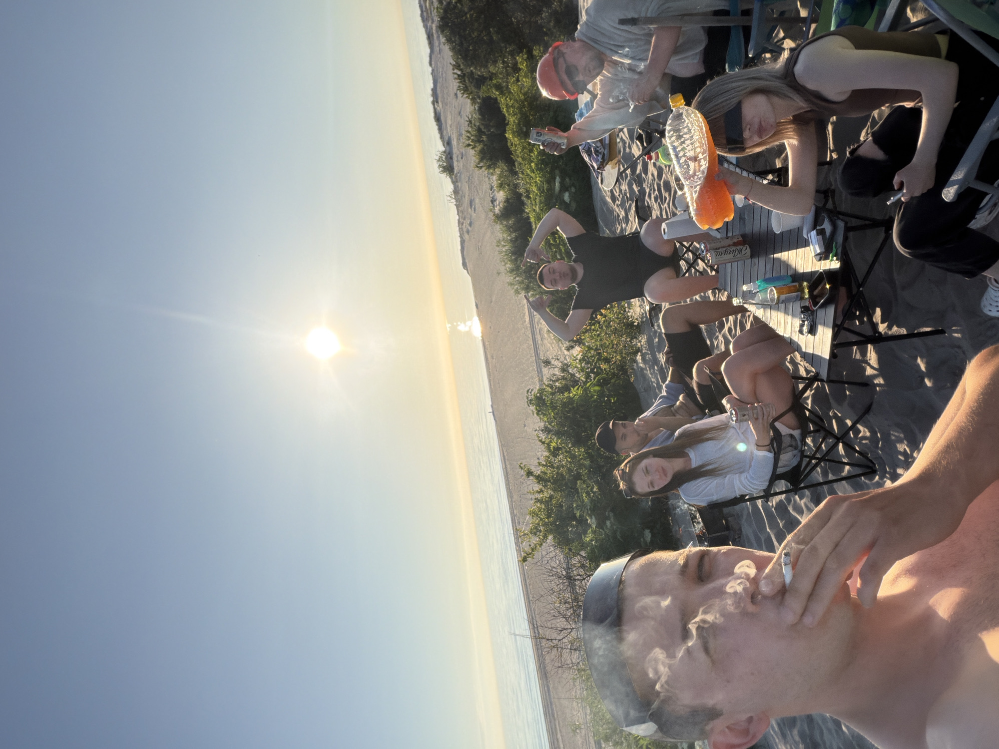 The image depicts a group of young adults gathered on a sandy beach near the city of Povarovka, Russia. The sun is setting over the calm sea, casting a warm glow on the scene. In the foreground, a young man is captured in a close-up, smoking a cigarette with smoke billowing around his face. He is wearing a black visor and appears to be topless.
Behind him, seated in folding chairs on the sand, are several other individuals. A young woman with long brown hair is looking towards the camera, holding a can. Next to her, a man in a dark t-shirt and shorts is seated, with his arms raised in a gesture. Further back, another man is visible, wearing a red cap and sunglasses, holding up a can and a bottle. A table is set up between them, holding various drinks and other items. The background shows the beach and vegetation, with the expanse of the sea and sky. The overall atmosphere appears casual and relaxed, suggesting a social gathering during sunset.