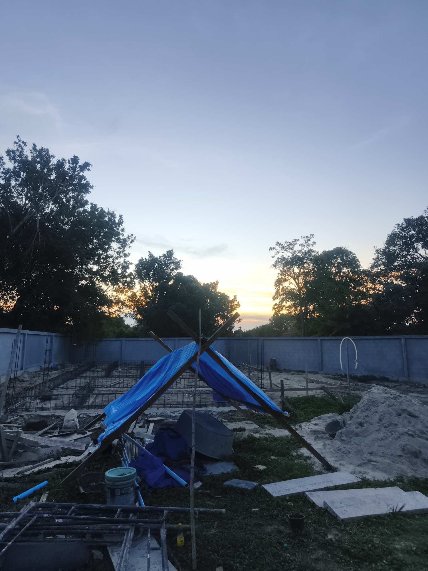 The image shows an outdoor construction site in Baan Mae Nam, Thailand at twilight. A blue tarp tent is erected on the ground among construction materials including rebar, lumber, concrete slabs, and plastic pipes. A wall surrounds the site, and trees line the horizon where the setting sun is visible.