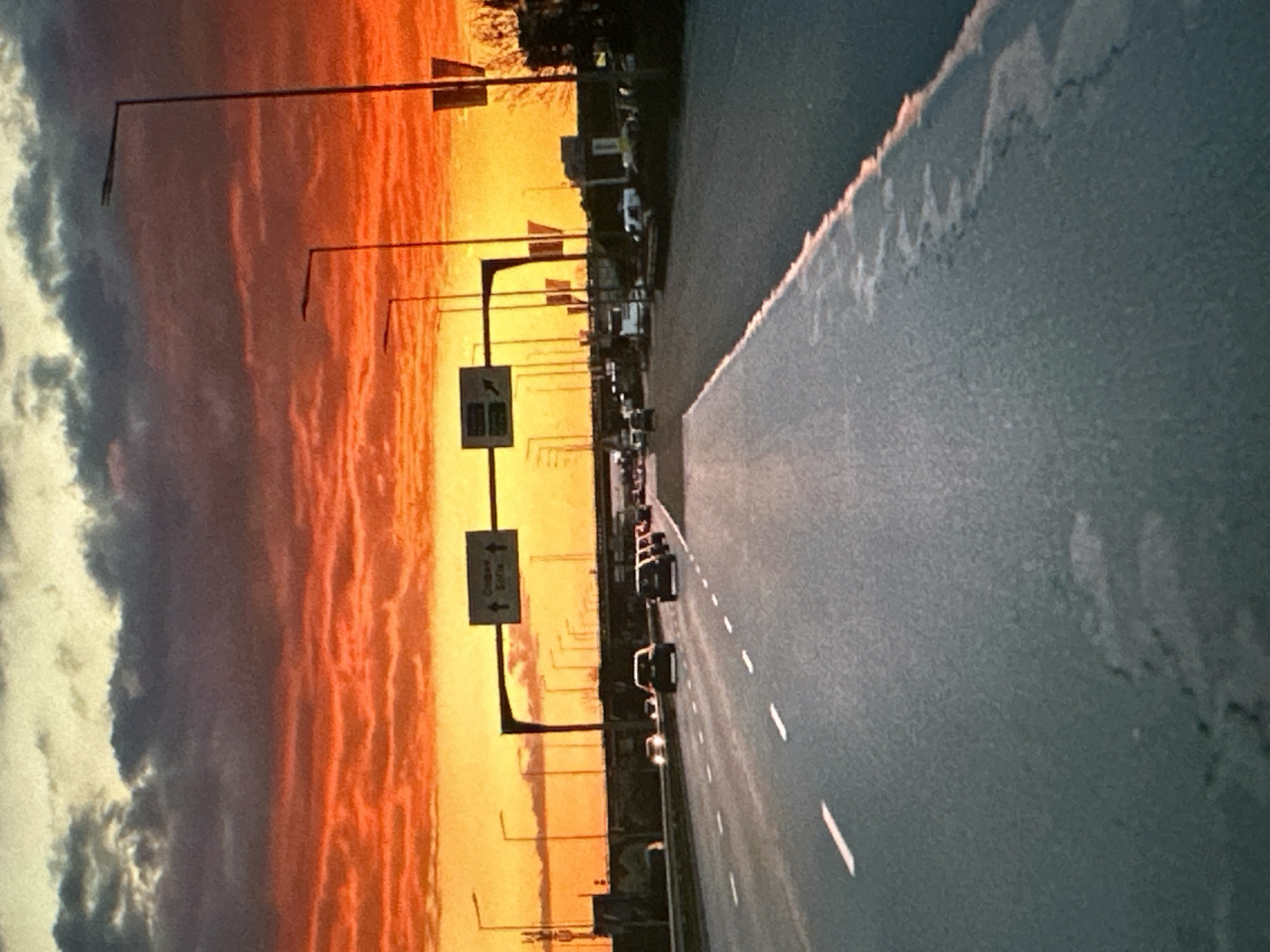 A street in Sofia, Bulgaria at sunset. The sky shows vibrant orange and yellow hues, transitioning into clouds. A road runs beside the street, with multiple cars. A signpost is present, pointing directions.