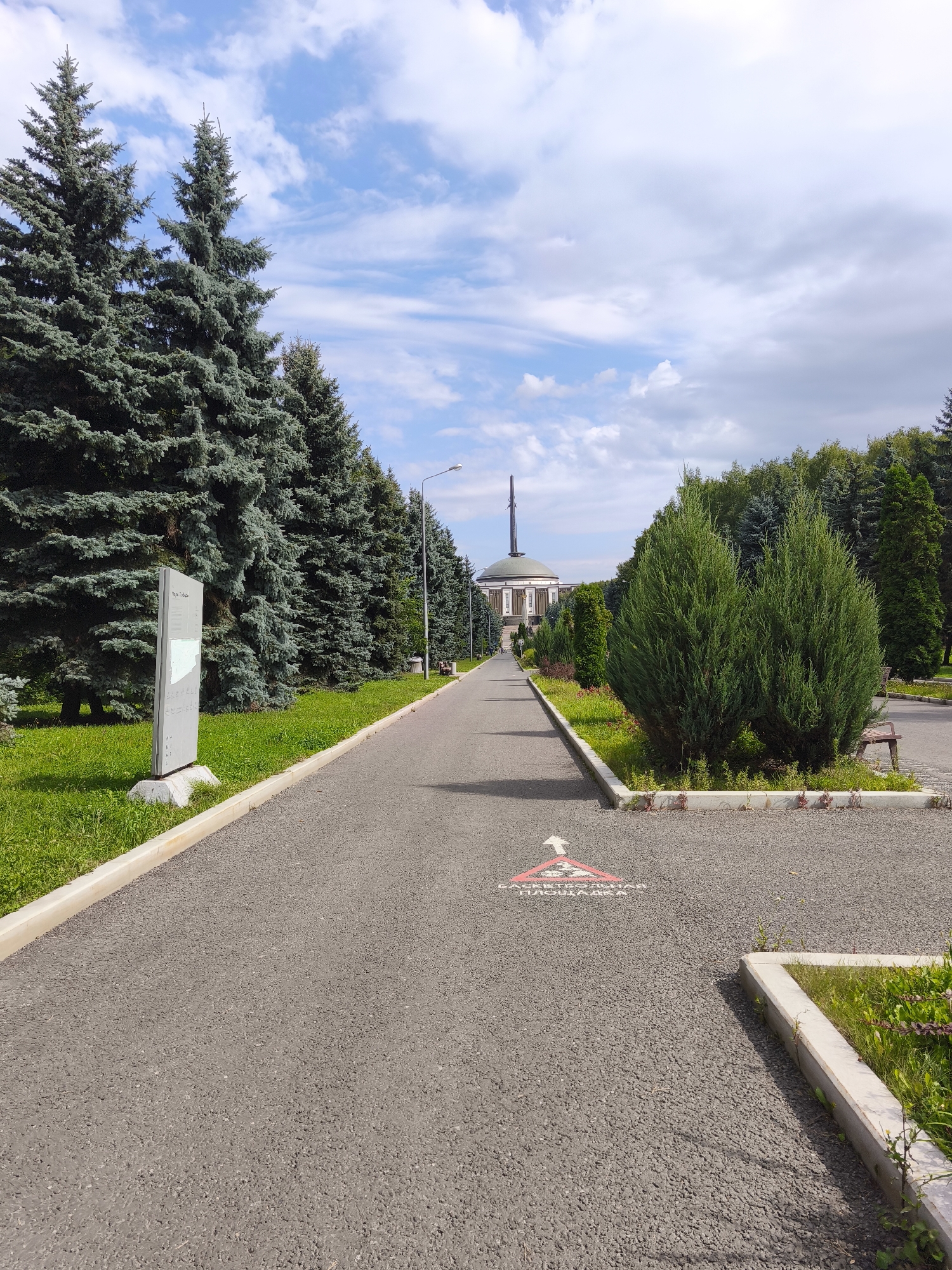 The image depicts a pathway leading towards a large, domed building with a tall spire, situated in a park-like setting with numerous evergreen trees. The pathway is paved with asphalt and bordered by grass and decorative landscaping. On the left side of the pathway, there is a tall informational sign. On the right side, there are also landscaped areas and a park bench visible in the distance. A small sign with an arrow and text is painted on the asphalt. The sky is blue with scattered white clouds. This scene is characteristic of the Great Patriotic War Central Museum (Central Museum of the Great Patriotic War) located at Poklonnaya Hill in Moscow, Russia.