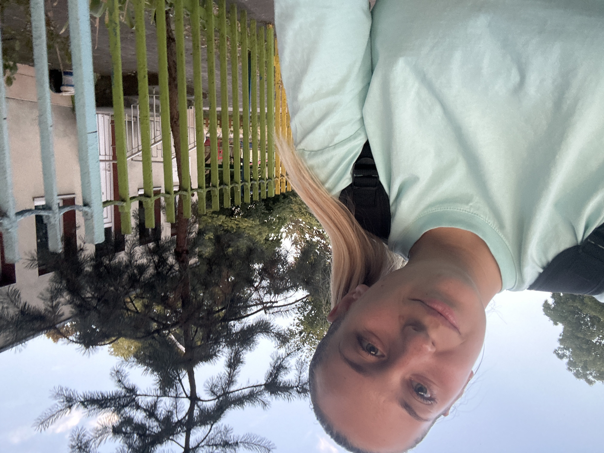A young woman with blonde hair pulled back in a ponytail is taking a selfie outdoors. She is wearing a light blue t-shirt and a black backpack. Behind her, there is a green and yellow fence, a tree with dark green needles, and a building with white walls and maroon accents. The sky is a pale blue with some clouds. The scene appears to be in a residential area, possibly a schoolyard or a park, in Plovdiv, Bulgaria.