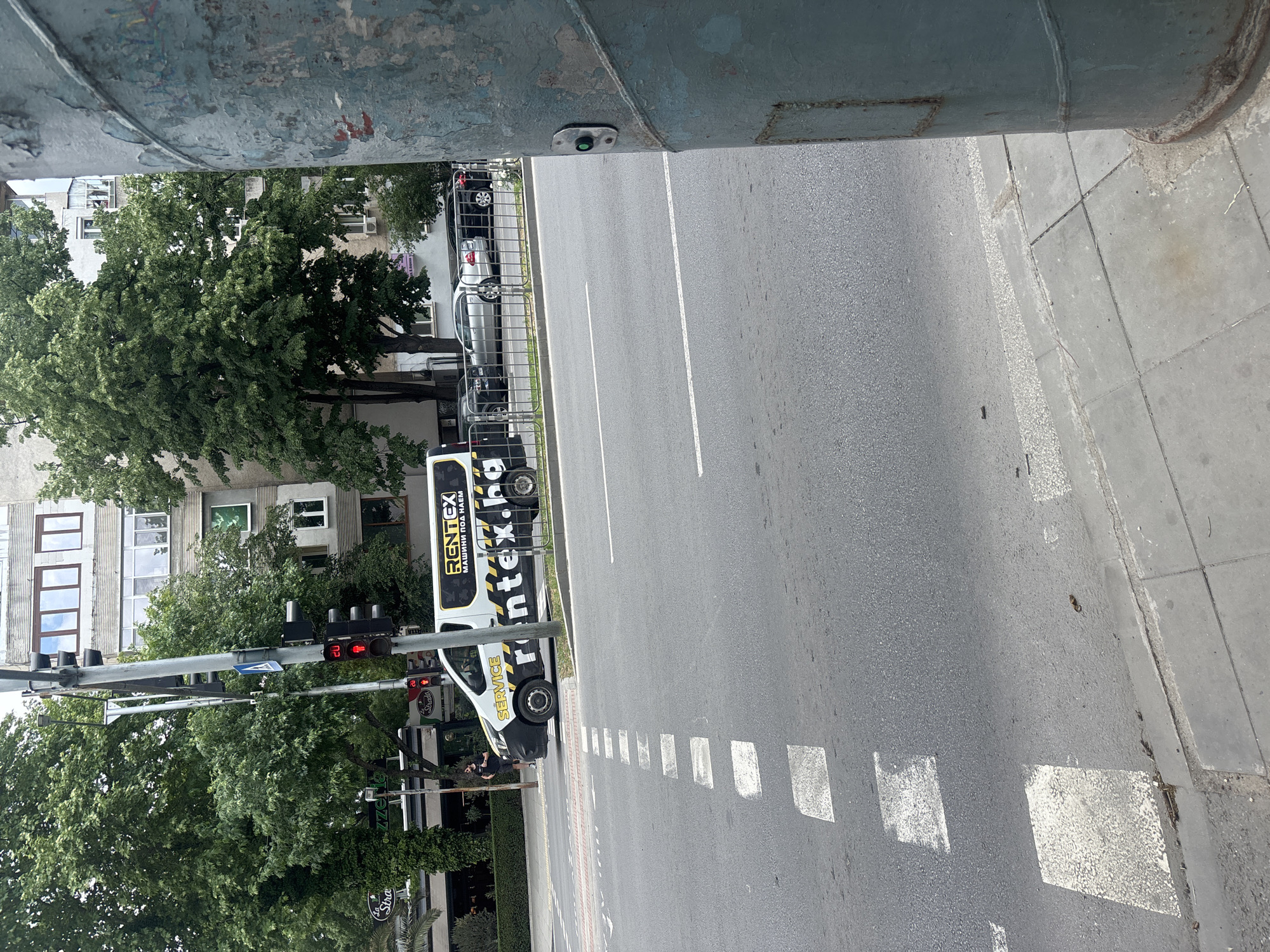 The image shows a street scene in Plovdiv, Bulgaria. A utility vehicle with "RENTEX" and a phone number is parked on the side of the road. A pedestrian stands near the vehicle. A crosswalk is marked on the road, with white squares painted on the asphalt. A traffic light displays a red signal. Trees and a multi-story building are visible in the background. The photo is taken from an elevated perspective, partially obscured by a pole. The day appears sunny and clear.