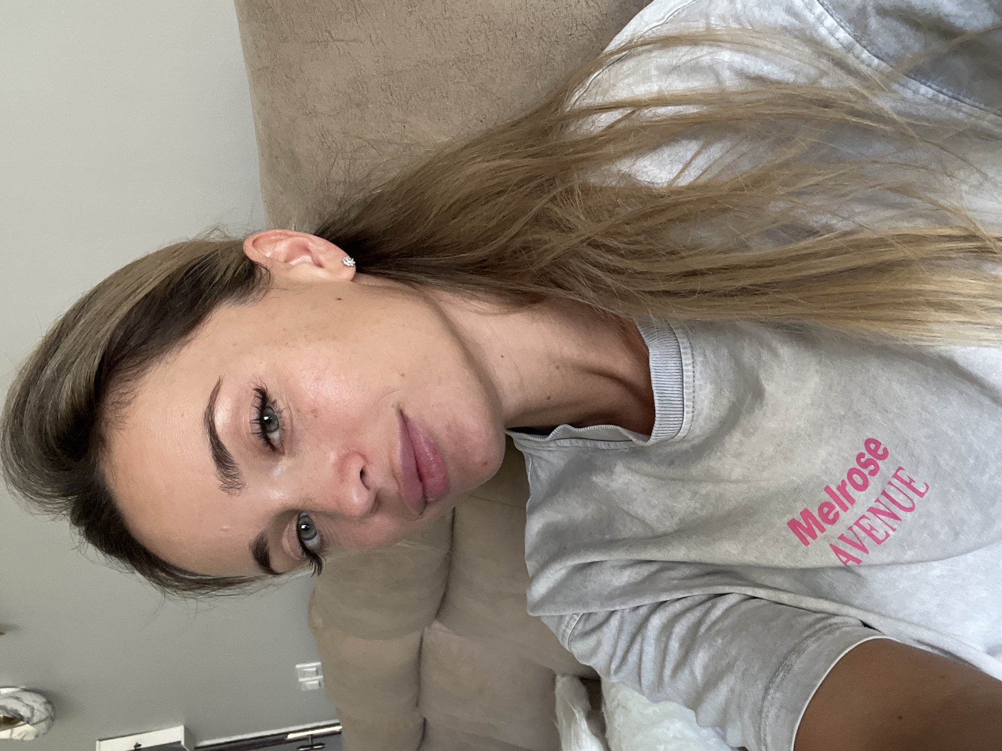 A young woman with blonde hair and green eyes is pictured in a selfie. She is wearing a grey t-shirt with "Melrose AVENUE" printed in pink on the front. She has on a small stud earring in her right ear. The woman is indoors, sitting on a couch, with her hair falling over her shoulder. The background is a neutral-colored wall and couch. No specific location in Sharm El Sheikh, Egypt, can be determined from the image.