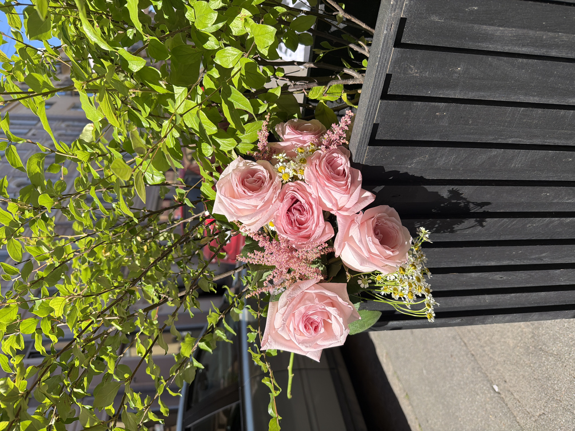 A bouquet of light pink roses with smaller pink flowers and daisies is arranged in a dark wooden planter box, which is situated outdoors on a sunny day. The planter is positioned in front of a lush green bush. In the background, a grey car is partially visible, and behind it, a building with windows can be seen. The scene appears to be in an urban setting, possibly in Saint Petersburg, Russia, given the architectural style of the building and the presence of a car and pavement. There are no people visible in the image.
