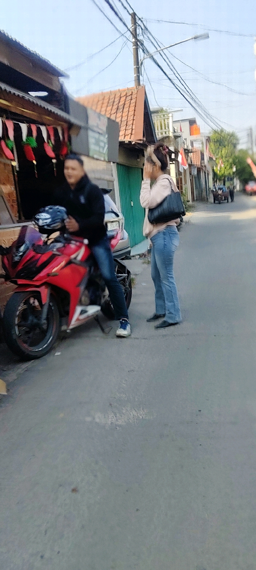 In Mekarmukti, Indonesia, a man is seated on a red motorcycle parked on the side of a paved road. He is wearing a black jacket and blue jeans. A black helmet rests on the handlebars of the motorcycle. To his right, a woman stands with her back to the viewer, looking away from the camera. She is wearing a light-colored hooded jacket and blue jeans, and carrying a black handbag over her shoulder. She has her hand raised to her face. Behind them, a building with a tiled roof is visible, and further down the street, other buildings and trees line the road. Power lines are strung across the sky.