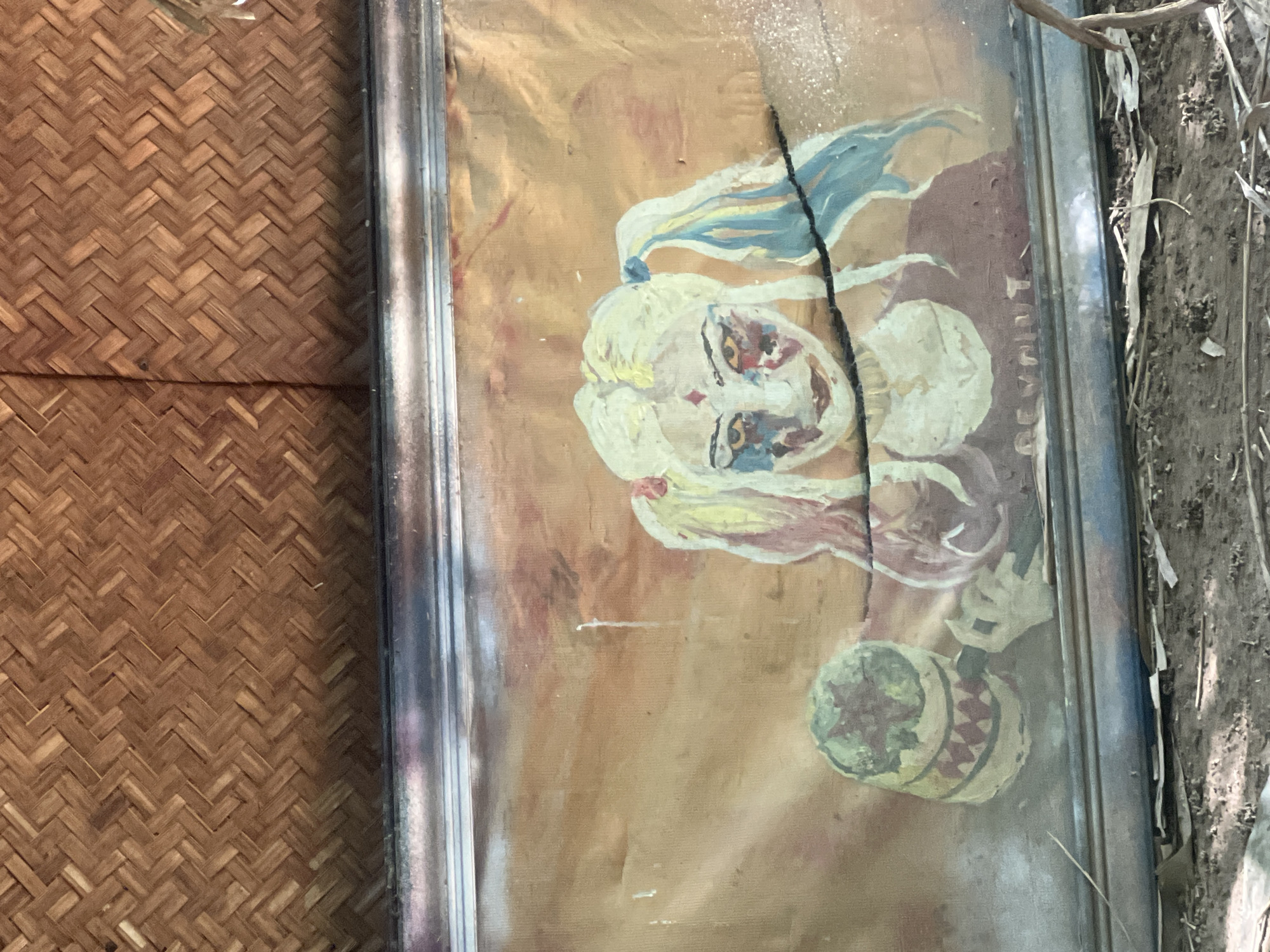 A framed painting of Harley Quinn is leaning against a woven bamboo wall. The painting depicts Harley Quinn with blonde pigtails, colorful makeup, and holding a mallet. The painting is cracked, and the frame is metallic. The background of the painting is a gradient of orange and brown. The environment appears to be an outdoor or abandoned setting, with dirt and debris on the ground. The location is not specified in the image or crops.
