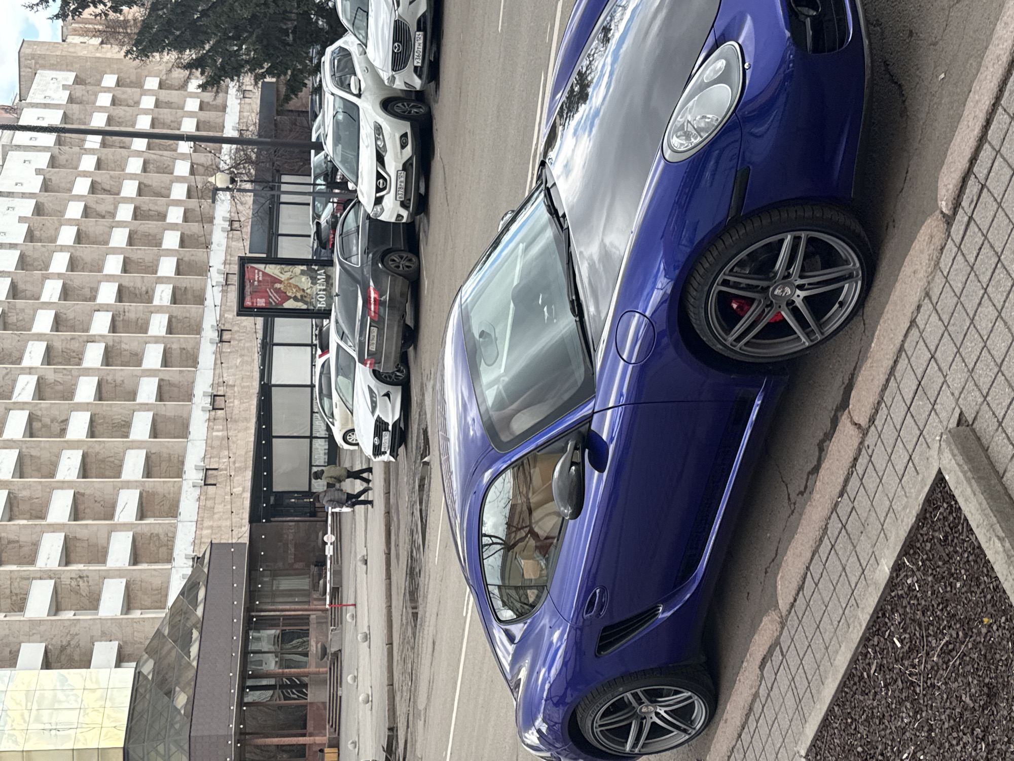 The image captures an urban street scene in Krasnoyarsk, Russia, featuring a striking blue sports car parked on the side of a road, next to a sidewalk and a row of other vehicles. The perspective is tilted, with the road running diagonally from the top left to the bottom right.
The central subject is a modified blue sports car, possibly a Porsche, with a dark (likely carbon fiber) hood. It has multi-spoke silver alloy wheels with red brake calipers visible behind them. The car's sleek design and custom features stand out.
Behind the blue sports car, a line of other vehicles is parked. These include several white SUVs, such as a Mazda CX-5 (license plate "360HC 124") and a Nissan Qashqai/X-Trail (license plate "870PB 124"), and a dark-colored sedan (possibly a Mercedes E-Class, with "706AA 124" partially visible on its license plate). The license plates indicate the region code "124," corresponding to Krasnoyarsk Krai.
On the left side of the frame, a large, multi-story building with a distinctive facade of numerous rectangular cut-outs or balconies dominates the background. Below this building, there's a more modern structure with glass and metallic elements, and a tall, dark vertical pole runs in front of it. Further in the background, a few bare trees are visible, suggesting it might be late autumn, winter, or early spring.
The time of day appears to be daytime, under an overcast sky, as reflected in the car's paint and the lack of harsh shadows. Two individuals are seen walking in the mid-ground, near a white and red barrier, suggesting a pedestrian crossing or entrance to a parking area. A rectangular sign with text is visible on a pole, displaying "БОГЕМА" (BOGEMA), which means "Bohemia" in Russian, and features an artistic image. The blue car's tire sidewall also shows "TIRELLES". The sidewalk features tiled paving, and a small planter with dark mulch is seen in the bottom right corner.