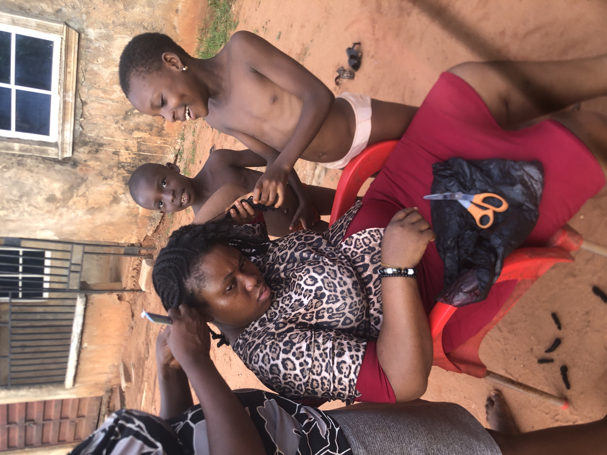 An outdoor scene in Ukwu-Abwa, Nigeria, captures a hair care activity involving several individuals on a reddish-brown dirt ground. The image is oriented sideways, but depicts a woman seated on a red plastic chair, wearing a leopard print top and a red skirt. Her hair is braided, and another person, mostly visible by their hands and arms wearing a black and white patterned top, is actively working on her hair. The seated woman has a neutral to slightly serious expression and wears a beaded bracelet on her left wrist.
Beside the seated woman, to her right, two young boys stand. The older boy, shirtless and wearing white briefs, smiles broadly with dimples and has a small gold earring in his left ear. He looks down at the hair activity. The younger boy, also shirtless, looks directly at the camera with a slight smile and holds a dark, fibrous object, possibly hair extensions or braids, in his hands.
On the seated woman's lap, a pair of orange-handled scissors rests on a black plastic bag. Scattered on the ground around the chair are numerous small, dark, cylindrical pieces, likely cut-off braids or hair extensions. A thin stick is also visible on the ground. In the background, a weathered building wall with a white-framed window is visible, along with a metal gate or fence structure further behind. The time of day appears to be daytime, under natural light. There is no discernible text in the image.