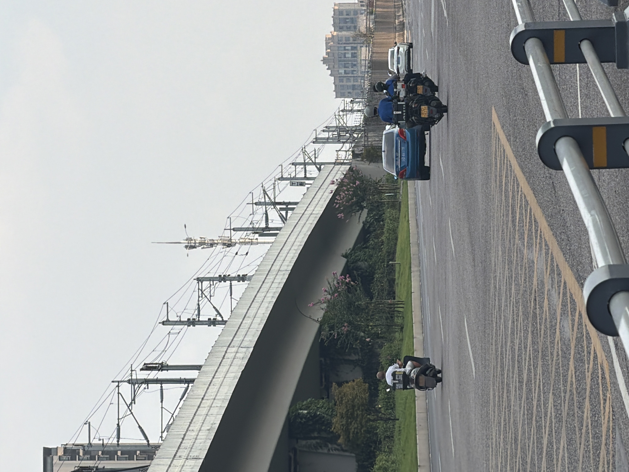 An elevated roadway, likely part of a highway or a significant urban artery in Jiangjin, China, dominates the upper left of the frame. The concrete structure spans across the scene, supported by large pillars. Overhead, a complex network of electrical wires and support poles for an unknown transportation system, possibly a light rail or high-speed train, crisscrosses the sky.
On the road below, a man on a white scooter, wearing a white shirt, rides away from the viewer. Further down the road, two individuals on a dark-colored motorcycle, both wearing helmets and dark clothing, are also traveling away from the camera. The motorcycle is laden with storage boxes. To the right of the motorcycle, a blue sedan is visible, and further still, a white sedan.
The road surface is asphalt with visible lane markings. A strip of green grass and some trees, including one with pink flowers, line the side of the road beneath the elevated structure. In the distance, a cluster of buildings, appearing to be commercial or residential, rises against the hazy sky. A tall communication tower with antennas is also visible on the right side of the frame. The overall impression is a busy urban environment with substantial infrastructure.