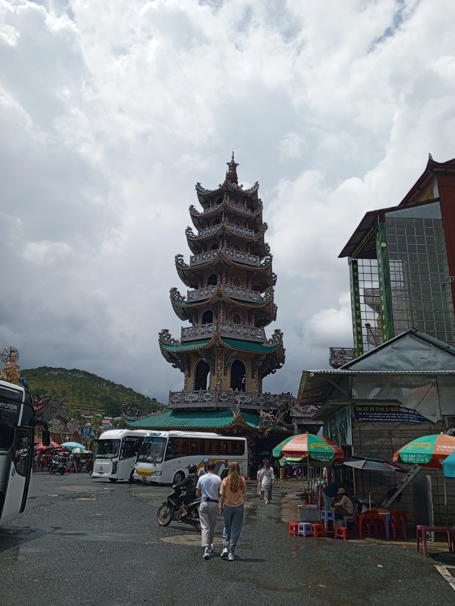 A tall, multi-tiered pagoda with ornate decorations and green tiled roofs dominates the center of the image. To the right, a modern building with large glass windows and a red brick facade stands adjacent to the pagoda. In the foreground, a paved area, wet with recent rain, shows several white tour buses and a motorcycle. Two individuals, a man and a woman, are walking away from the camera on the wet pavement, while another person rides a motorcycle. Several small stalls with umbrellas are set up on the right side of the scene. In the background, a lush green hillside is visible beneath a cloudy sky. The scene appears to be a public area, likely a tourist attraction or transportation hub in Xuân Thành, Vietnam.