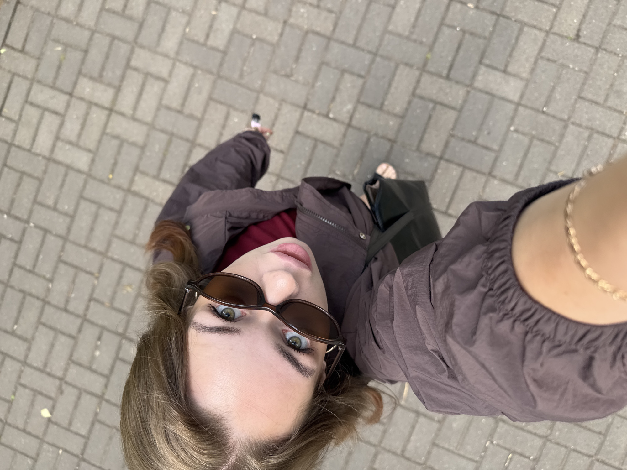 A high-angle, close-up, selfie-style shot shows a young woman with light brown hair and green eyes looking up at the camera. She is wearing dark brown sunglasses and a dark brown jacket over a red top. Her left arm is extended, holding what appears to be a smartphone to take the picture. A black handbag is visible on the ground next to her. The woman is standing on a grey brick-paved walkway in Chaikovsky, Russia. The walkway is made of rectangular bricks laid in a staggered pattern. There are a few scattered leaves on the ground. The lighting suggests it is daytime.