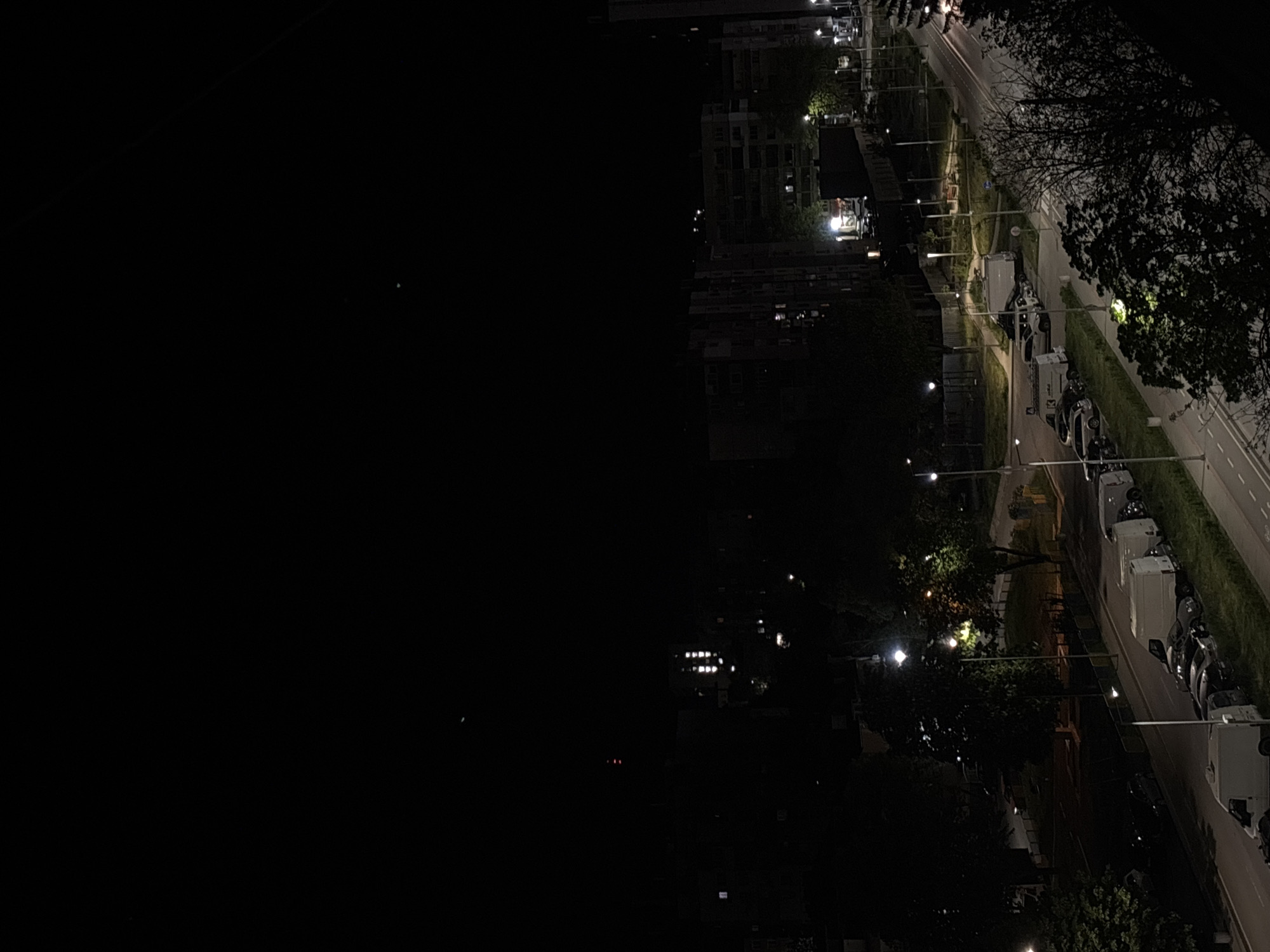 The image depicts an urban environment at night. A residential area with buildings is visible, illuminated by streetlights and apartment windows. A road runs alongside the buildings, with parked cars and trees lining the side. The sky is dark and featureless.