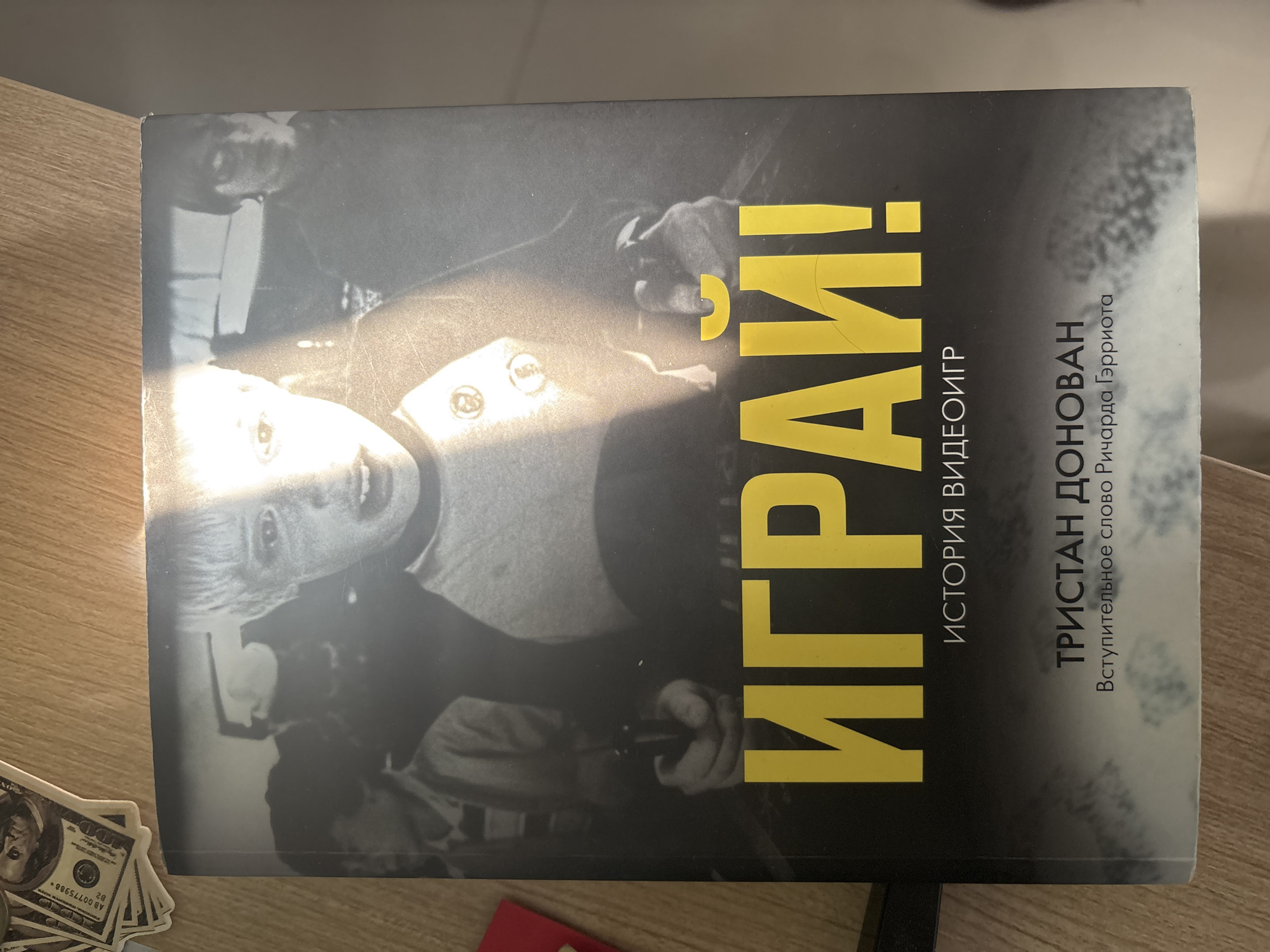 The image shows the cover of a book titled "ИГРАЙ!" which translates to "PLAY!". The cover features a black and white photograph of several people, with a young boy in the foreground looking directly at the camera with his mouth open. The text below the title reads "ИСТОРИЯ ВИДЕОИГР" which means "HISTORY OF VIDEO GAMES". Below this, the author's name is listed as "ТРИСТАН ДОНОВАН" (Tristan Donovan), and the introductory word is by "Ричарда Гэрриота" (Richard Garriott). The book is lying on a wooden surface, and a few dollar bills are visible in the upper left corner of the image. The scene is not a city, but rather a close-up of a book cover on a table.
