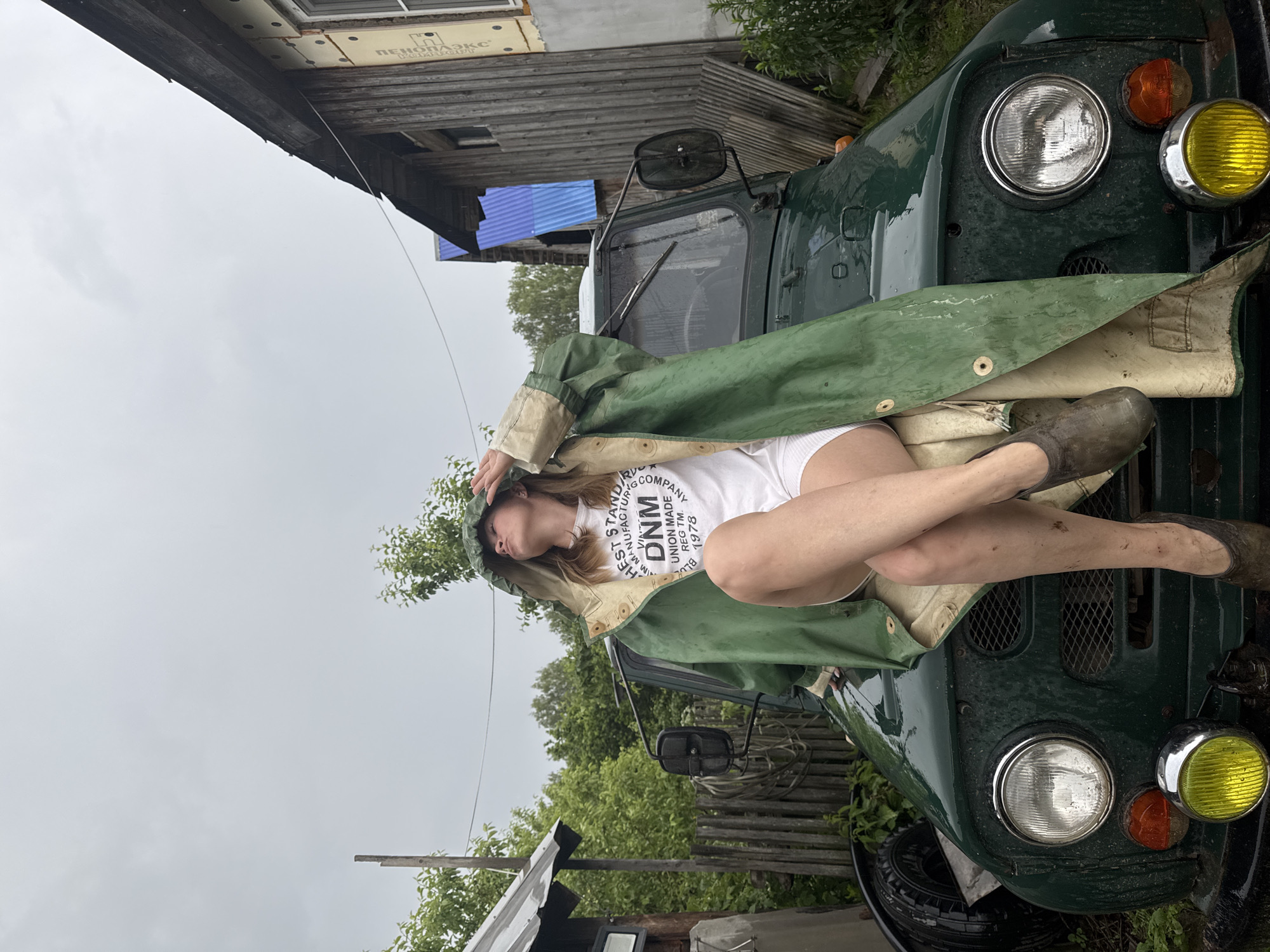 The image depicts a woman sitting on the hood of a green, utilitarian vehicle, likely an off-road vehicle, in what appears to be a rural or semi-rural setting. She is wearing a green raincoat, a white t-shirt with text, white shorts, and dark, muddy clogs. Her pose suggests a relaxed or contemplative demeanor, with one hand raised to her forehead as if shielding her eyes or adjusting her hood. The vehicle's headlights, grill, and fenders are visible. In the background, there are wooden structures, a fence, trees, and a cloudy, overcast sky, indicating potentially inclement weather. The overall scene suggests a simple, perhaps weathered, environment. The location is identified as Vas'yata, Russia, a village in the Amur Oblast.