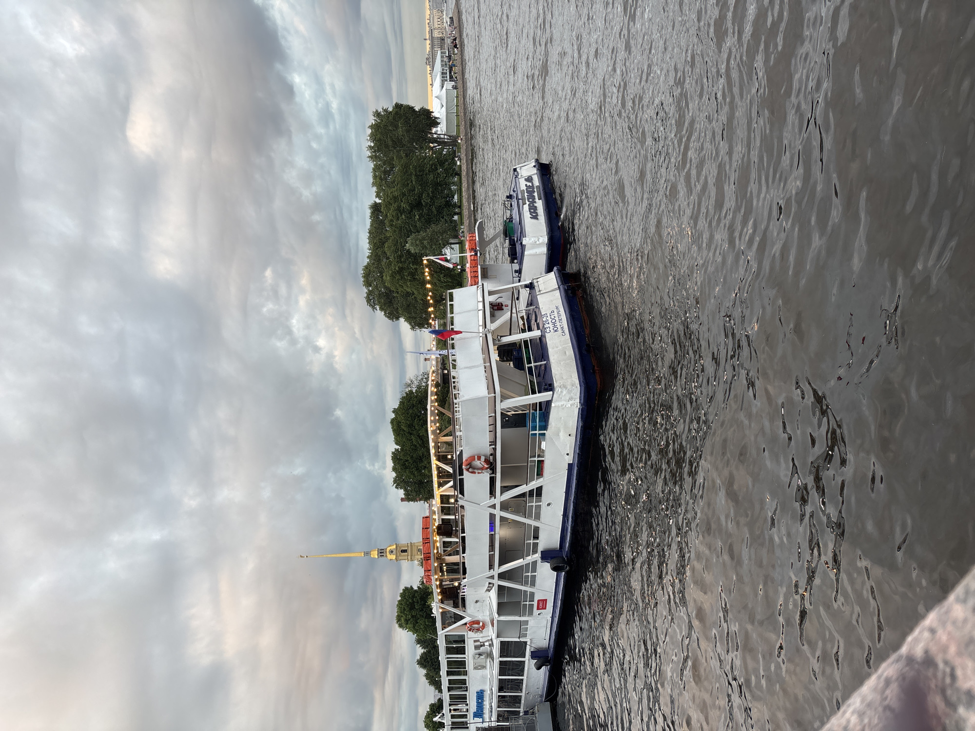 The image depicts a cloudy day at a river in Saint Petersburg, Russia. In the foreground, two white tour boats are docked. The larger boat on the left has "ИМПЕРИЯ" written on its side in blue lettering. The smaller boat on the right has "КОМЕТА" written on its stern. Behind the boats, trees line the riverbank, and a tall, slender spire topped with a golden dome and a flag rises above the trees. This spire is characteristic of the Peter and Paul Fortress, a prominent landmark in Saint Petersburg. Further in the background, several white tents are visible on the shore, and buildings can be seen in the distance. The water is dark and choppy, reflecting the overcast sky. There are no discernible people actively engaged in actions, though figures may be present on the distant shore. The overall scene suggests a tourist area where boat tours are a common activity.