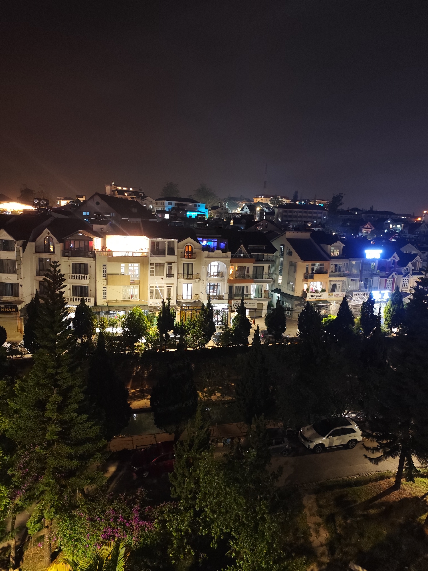 Captured at night, the image presents an elevated view of a section of Da Lat, Vietnam. The foreground contains a mix of trees and shrubbery, partially obscuring two parked vehicles: a red car and a white SUV. The middle ground features multi-story buildings with white facades and varied architectural styles, illuminated by a combination of interior lighting and streetlights. In the background, the cityscape extends into the night, with faint lights suggesting a broader urban area. The sky above is dark and featureless. No people are visible in the scene.