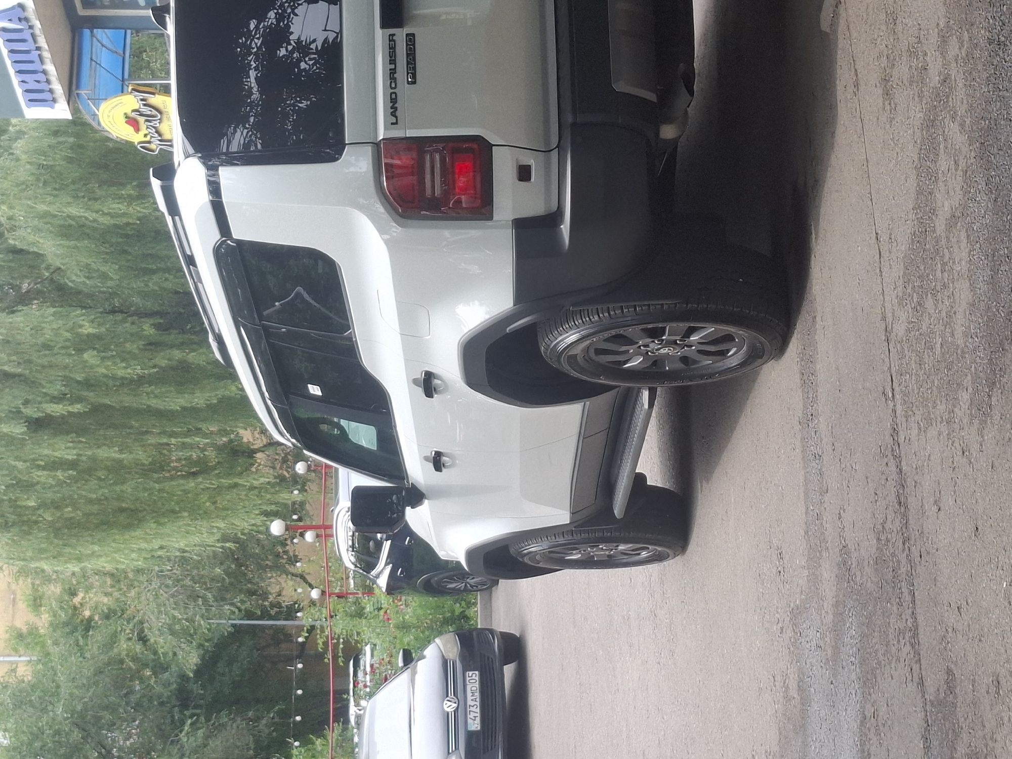 In Almaty, Kazakhstan, a white Toyota Land Cruiser Prado is parked on a paved lot. The vehicle's rear-left quarter is prominently featured, with the tail light illuminated. To the left of the Prado, a black Volkswagen Polo is parked, displaying a license plate that reads "KZ 473 AMD 05". In the background, a building with a sign that says "ПИЦЦА" (Pizza) is visible, along with trees and streetlights. The overall scene depicts an urban outdoor setting.