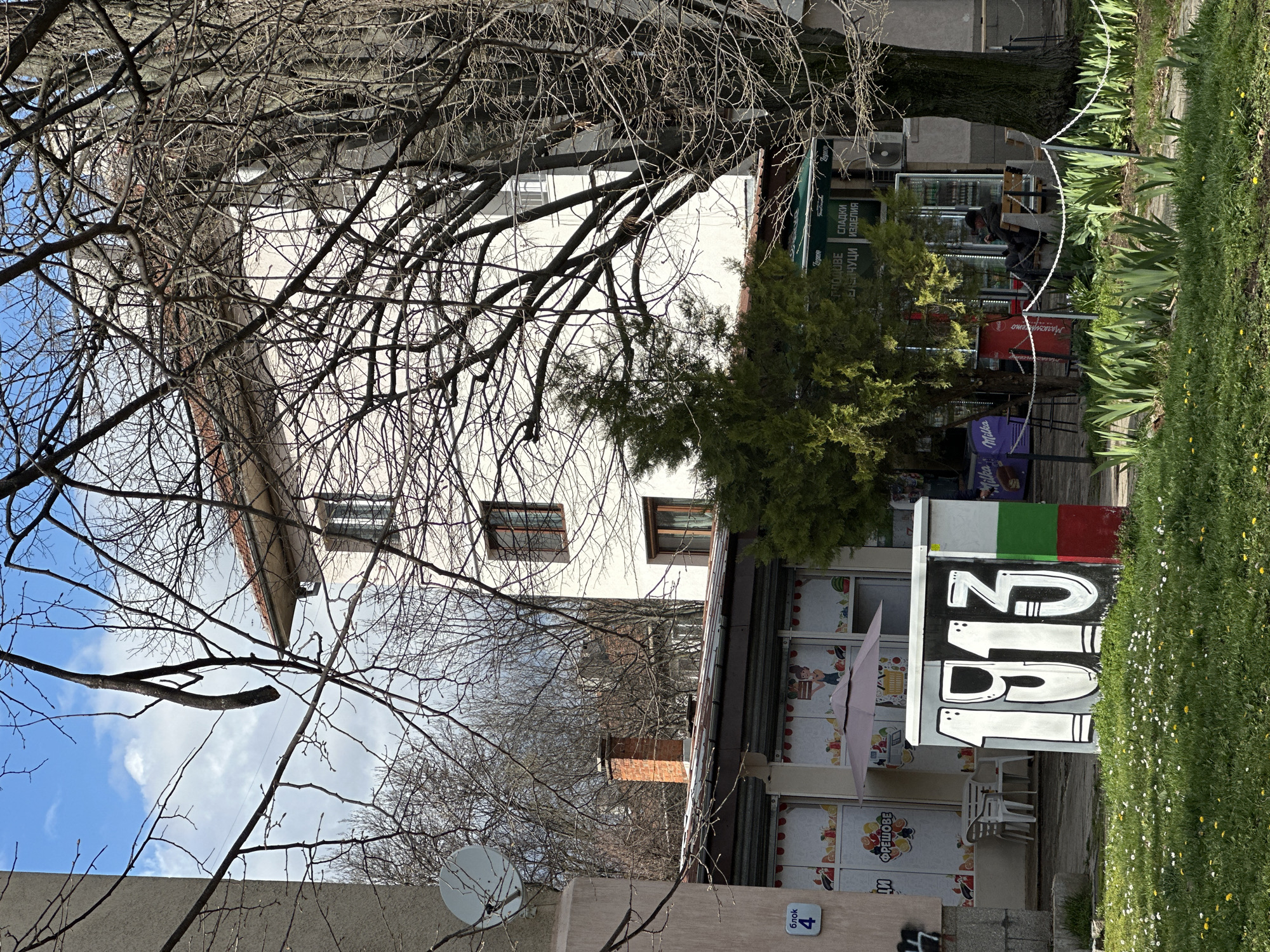 The image shows a street scene in Sofia, Bulgaria, featuring a small shop. The shop has large windows decorated with fruit and cartoon character decals, and a sign displays "153," with the Italian flag painted above. Outdoor seating is visible, with white plastic chairs and tables. A tree obscures part of the shop. Adjacent to the shop is a two-story white building with visible windows. In the foreground, a wall with the number "4" is visible, and bare tree branches frame the scene. Grass with small white and yellow flowers is present in the foreground. A person is seated near a storefront beyond the initial shop. A satellite dish is mounted on the wall on the left.