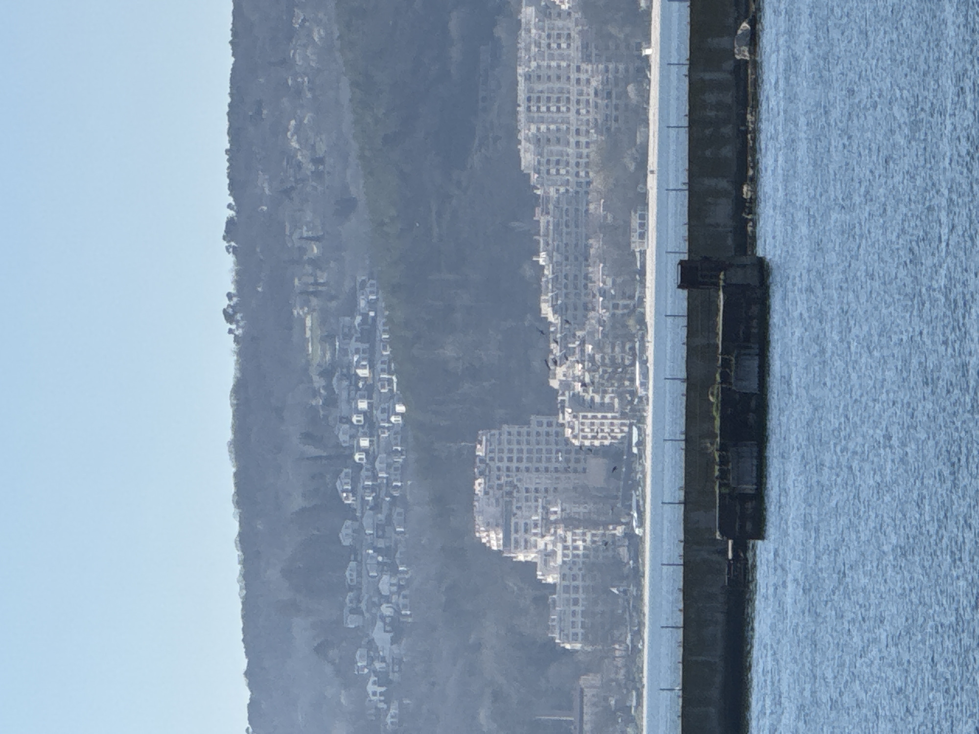 The image shows a view in Varna, Bulgaria, encompassing land, buildings, and water. On the left is a hillside with densely packed residential houses, followed by high-rise apartment buildings. To the right, a concrete pier separates the land from the sea. The water is calm with subtle texture and reflections. The sky is clear with a pale blue color.