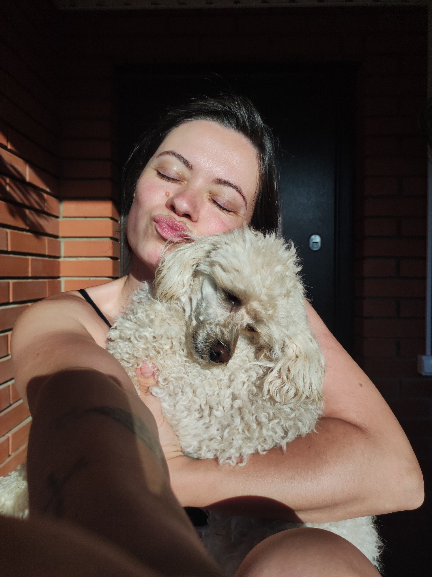 A woman is holding a white poodle in her arms. She has dark hair and is making a kissing face with her eyes closed. She has a mole on her face. Her left arm is partially obscured by a shadow. The background consists of a red brick wall and a black door. The photograph appears to have been taken in sunlight. The precise location in the city of Фоки, Russia cannot be determined from this image.
