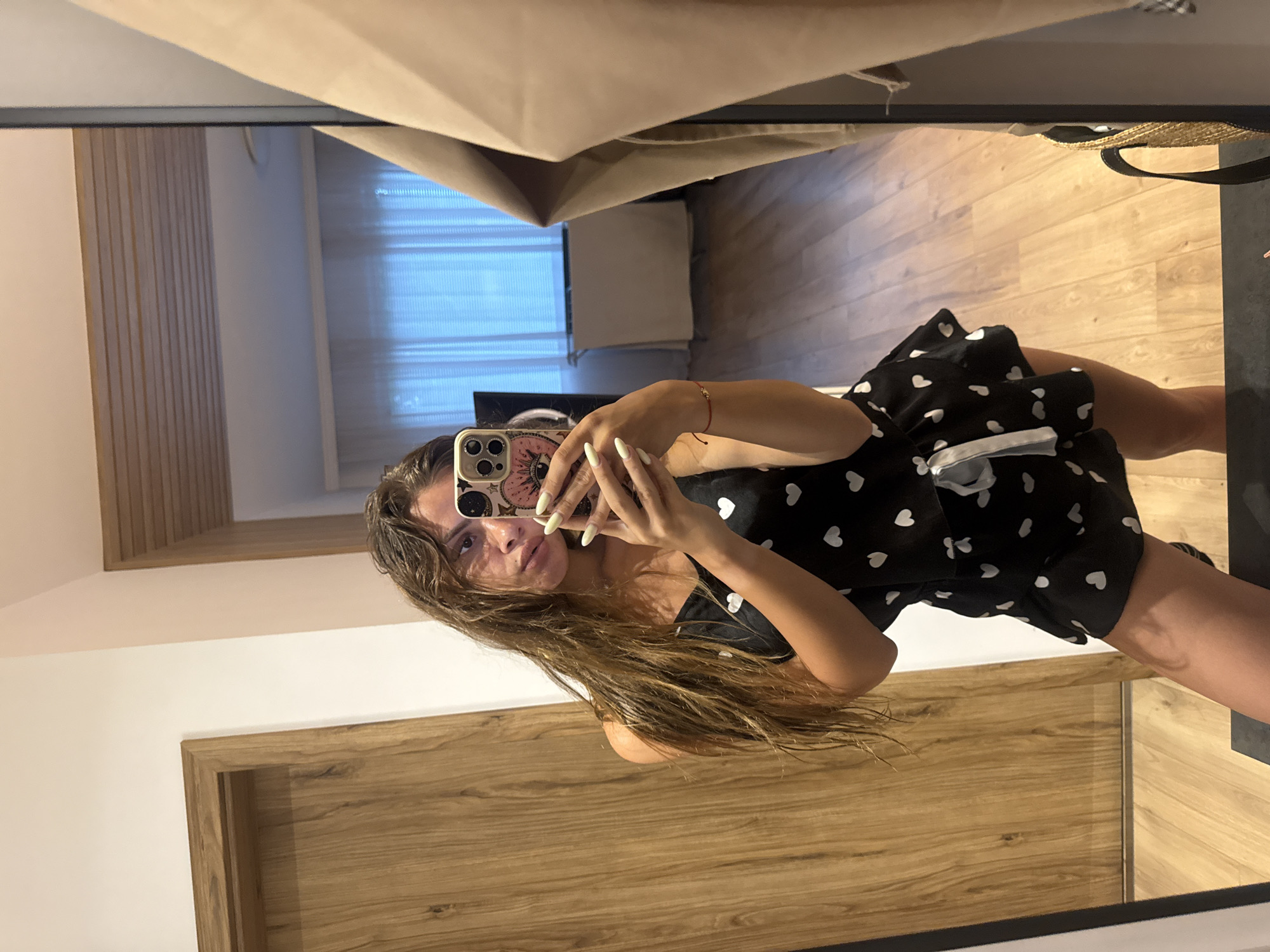 A young woman with long, dark, wavy hair takes a mirror selfie in what appears to be a modern apartment bedroom in Plovdiv, Bulgaria. She is wearing a black slip dress with a pattern of white hearts and a white bow. The room has light-colored wooden floors and a wooden door to her left. In the background, seen in the mirror, is a window with horizontal blue blinds and a beige-colored radiator. A large mirror dominates the right side of the frame, reflecting the woman and part of the room. She is holding a smartphone with a decorative case, and her nails are painted a light green color.