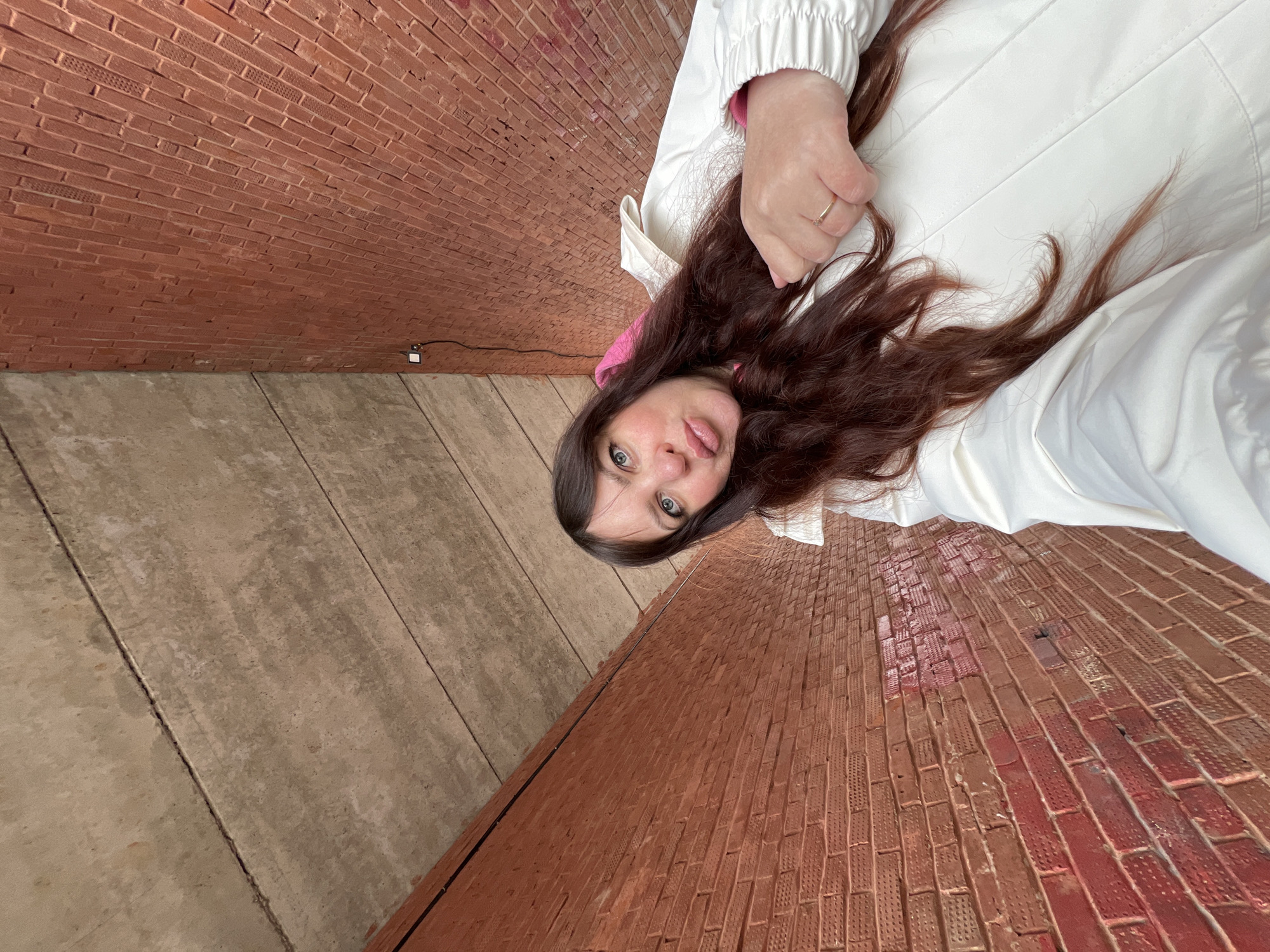 A woman with long brown hair, wearing a white jacket over a pink top, is taking a selfie. She is standing in an outdoor tunnel-like structure. The walls consist of red brick on one side and gray concrete slabs on the other. A small electrical box with wires is visible on the brick wall near the transition to the concrete. The photo is angled upward. It is unknown whether the location is Noviy, Russia.