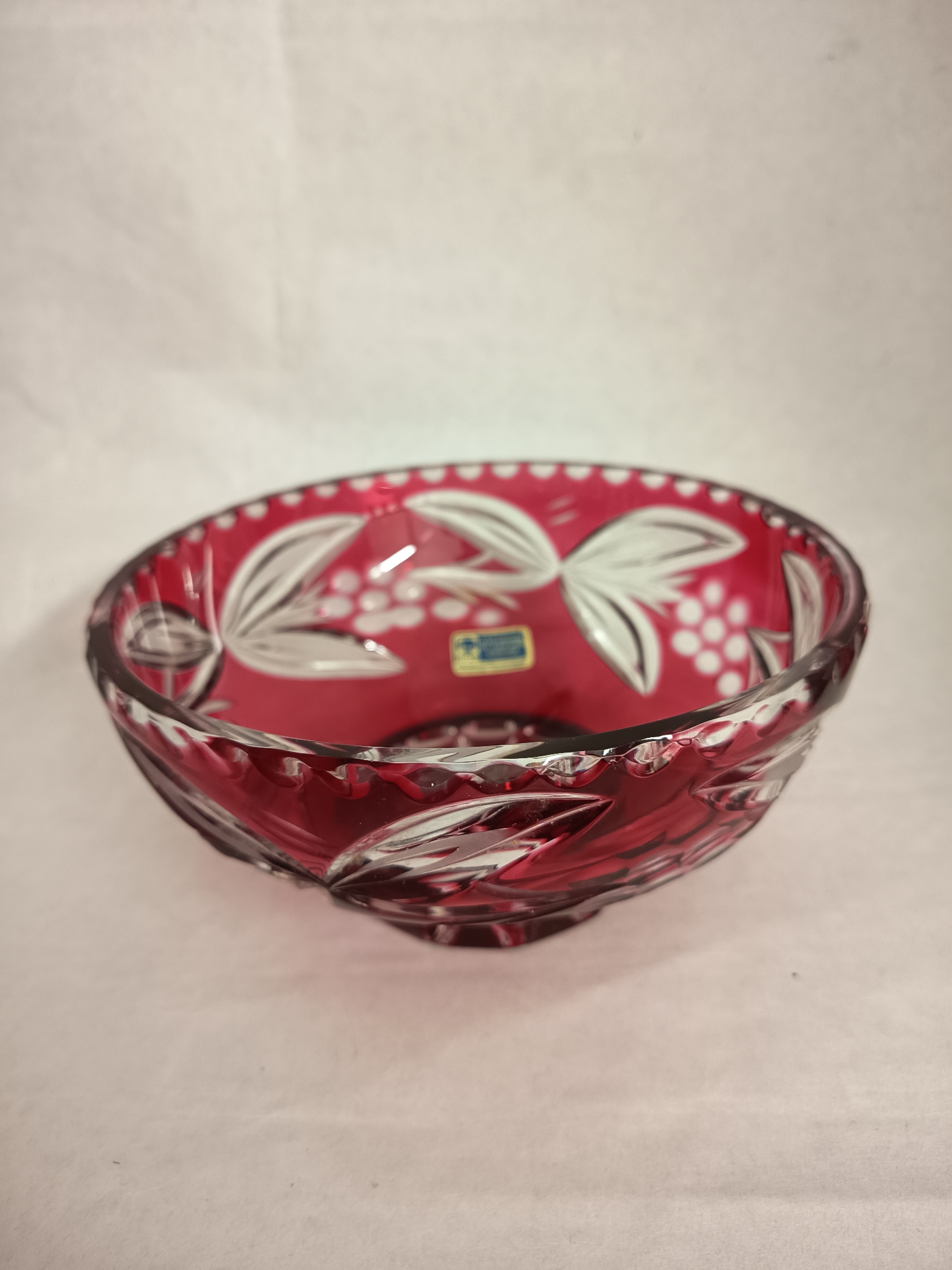 A red crystal bowl with white floral etchings is centered against a white background. A small blue and yellow sticker is affixed to the inside of the bowl. There are no people visible in the frame. The location of Wangen bei Olten, Switzerland is not identifiable within the image.