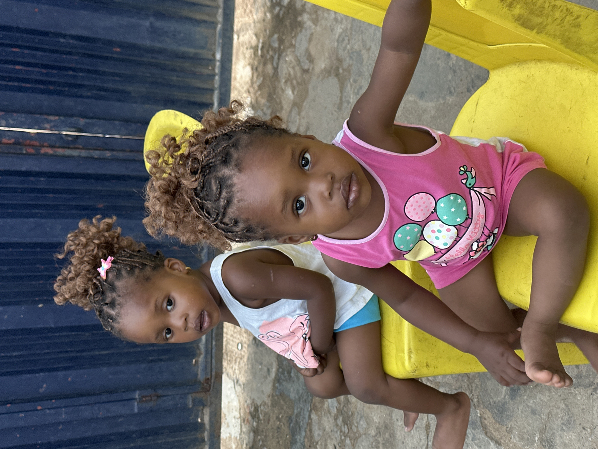 An outdoor, rotated image depicts two young children, likely girls, sitting on a yellow plastic play structure, possibly a slide or a chair, in Narayi, Nigeria. The children are positioned side-by-side, looking directly at the camera with neutral expressions. The child on the right wears a bright pink tank top adorned with colorful balloons and a character design, with "Hello Kitty" text visible on it. She has braided hair styled into several puffs. The child on the left wears a white tank top with a pink design and blue shorts, and her braided hair is styled into a single puff with a small pink clip.
The background consists of a dark blue, possibly corrugated metal, wall or fence, with hints of red-brown rust or streaks. The ground beneath the children is a light-colored concrete or paved surface. The lighting suggests it is daytime with clear weather. There are no other prominent activities occurring, the focus being on the children's direct gaze.