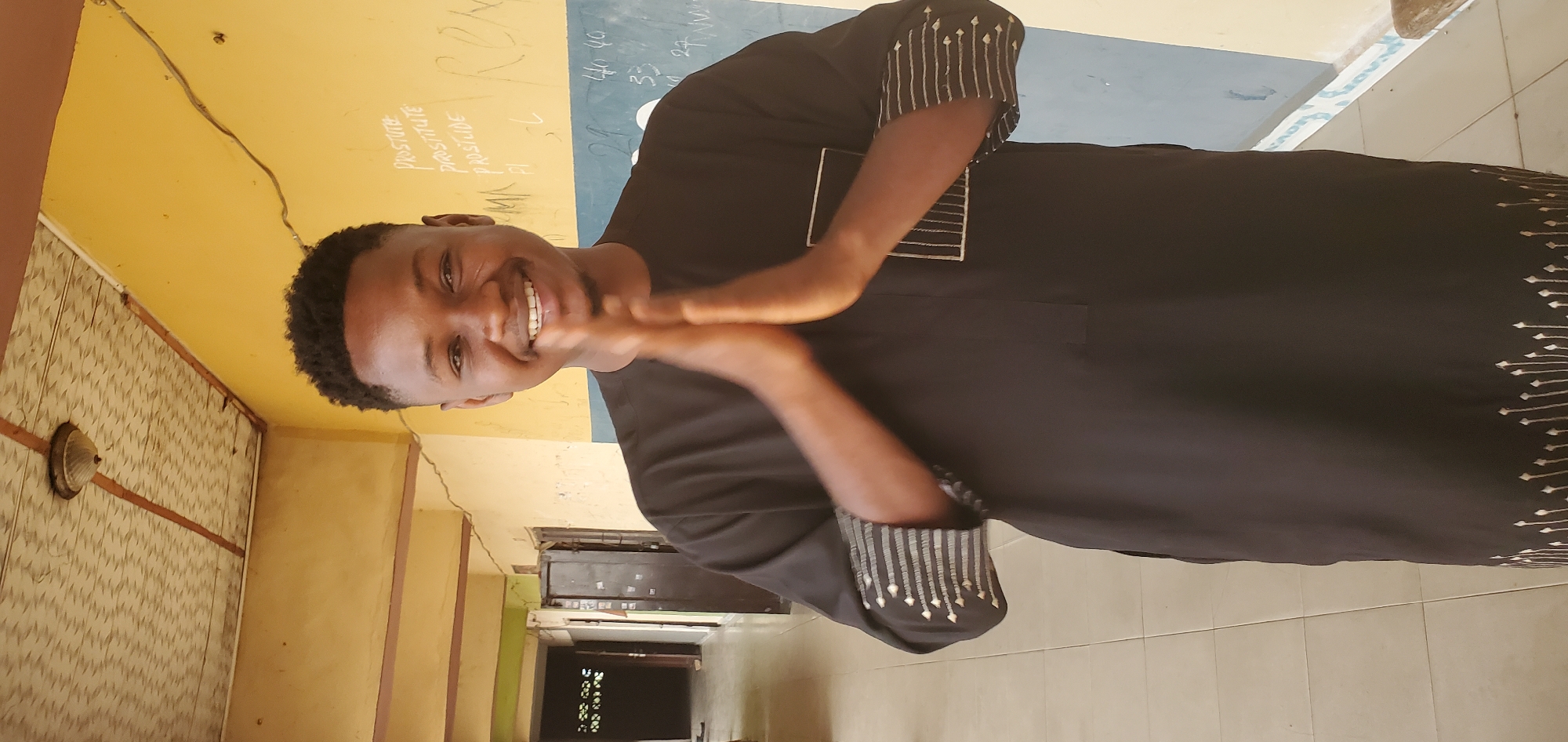 A man stands indoors, smiling, and clapping his hands together in Mararaba, Nigeria. He has a short haircut and wears a long-sleeved black garment with embroidered details on the sleeves and lower hem. The background consists of a yellow wall with writing and a blue chalkboard. A hallway stretches into the distance.