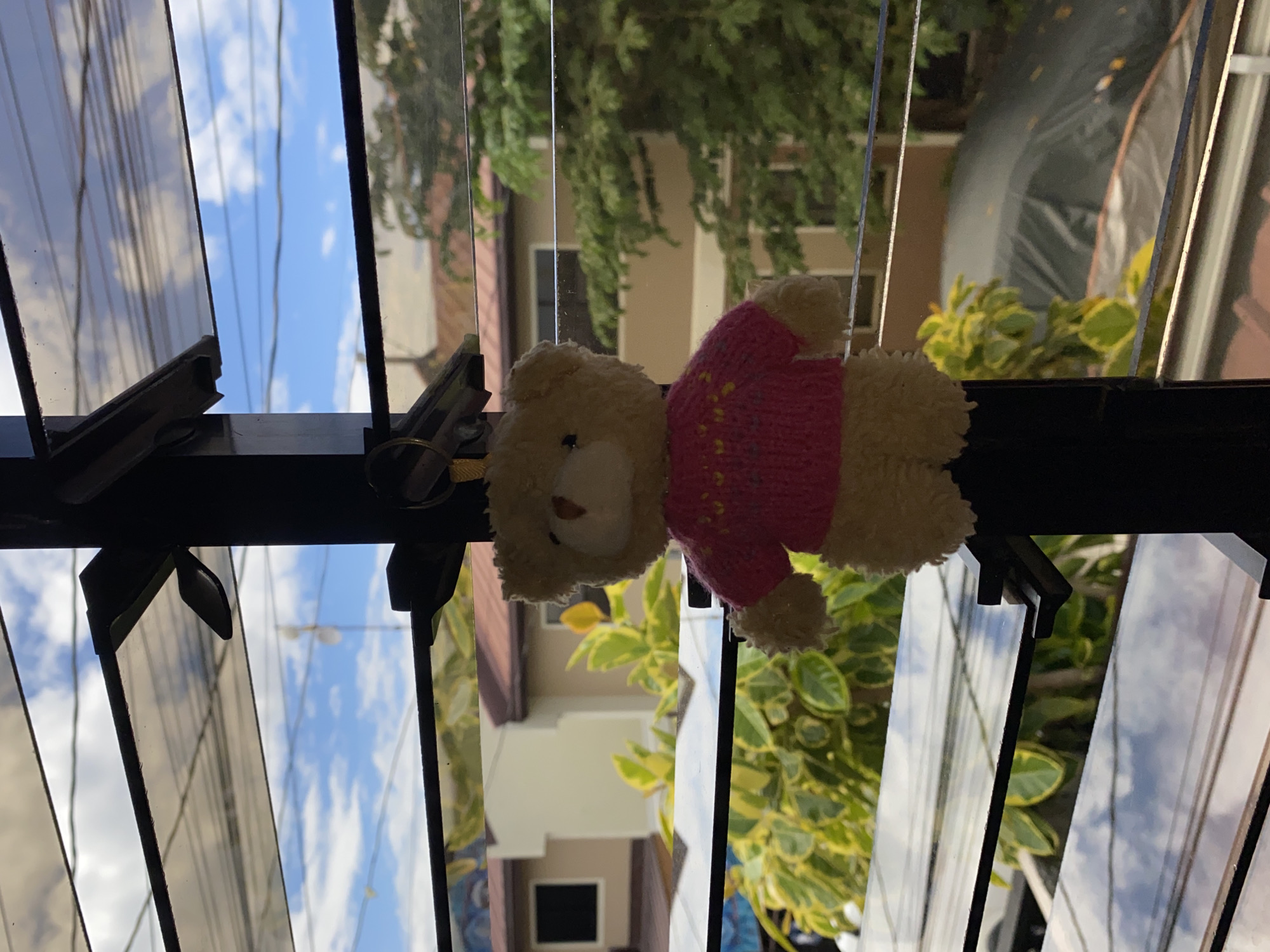 A beige teddy bear wearing a pink knitted sweater with yellow and white patterns hangs from a black metal frame in front of a jalousie window. The window overlooks a residential area in Davao City, Philippines, with houses and lush green trees visible under a bright, cloudy sky. The scene is framed by the horizontal slats of the jalousie window, which reflect the sky and clouds. The teddy bear is positioned centrally, with its body slightly tilted to the left. The background shows the typical architecture and vegetation of a tropical urban environment.