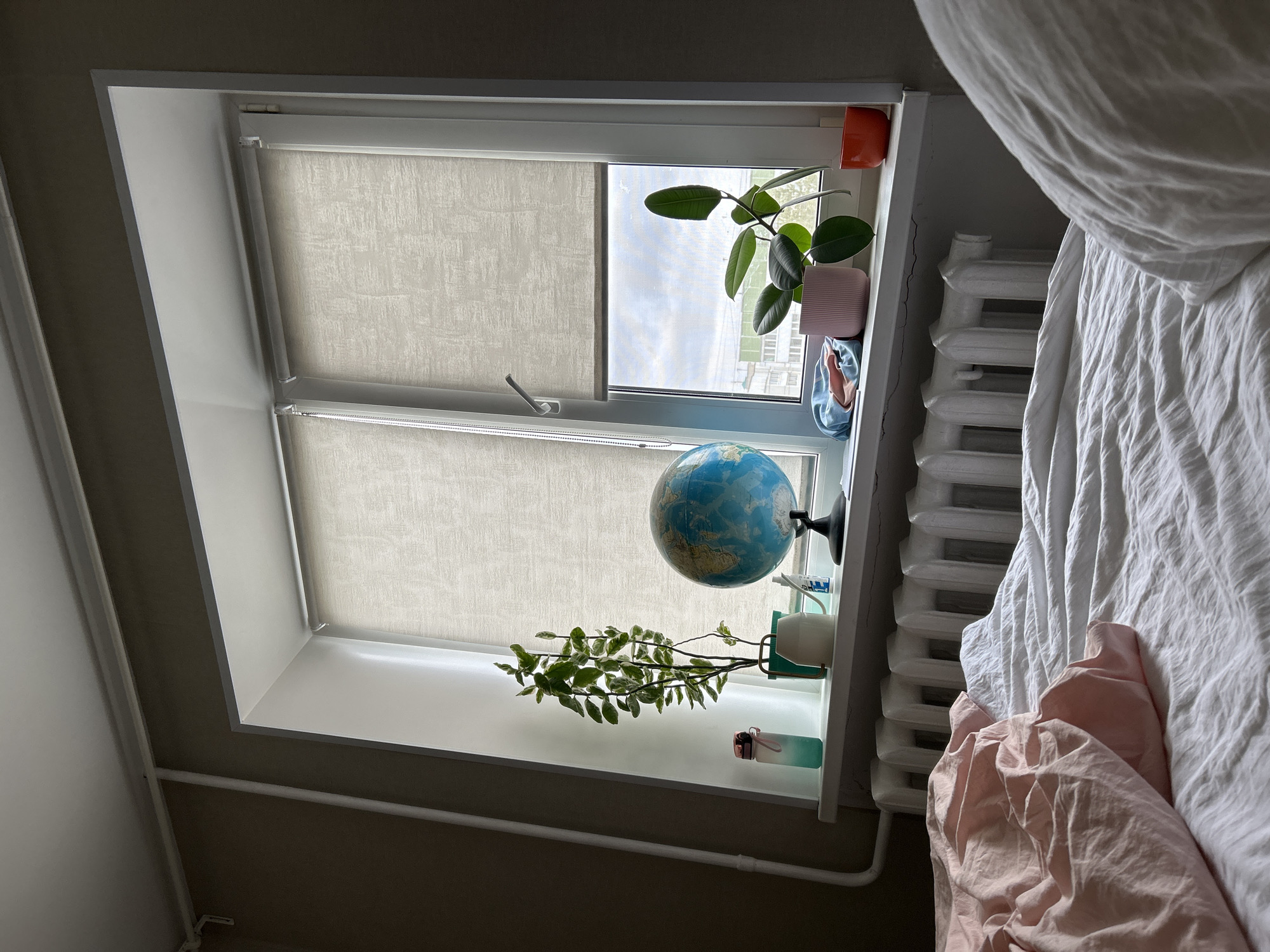 This is an interior, eye-level shot of a room, likely a bedroom, in Chaikovsky, Russia, focusing on a window area. The scene is static, depicting no activity.
The central subject is a wide window with a white frame and two partially drawn, textured beige roller blinds. Light filters in from outside, indicating daytime under an overcast or cloudy sky, with distant buildings visible through the window, suggesting an urban or suburban setting.
The window sills are adorned with various objects:
- On the upper sill to the right, a potted rubber plant (Ficus elastica) with large, dark green leaves sits in a pink pot. Beside it are an orange pot or mug, a small patterned dish, and a bundled piece of blue fabric.
- On the lower sill, a blue globe with a black stand is prominent. To its left is a trailing plant with variegated green and white leaves in a white pot. A white cup/mug and a small, translucent bottle with a pink cap are also present.
To the right of the window, a white, ribbed, cast-iron radiator is mounted on the wall. Above it, minor cracks are visible in the paintwork below the window sill. A portion of a bed is visible on the far right, covered with white bedding and a pink blanket or duvet. White pipes run along the lower part of the wall. The walls surrounding the window are light-colored, while other sections appear a darker, brownish-gray.
No visible text is discernible in the image. The overall atmosphere of the room appears lived-in and peaceful.