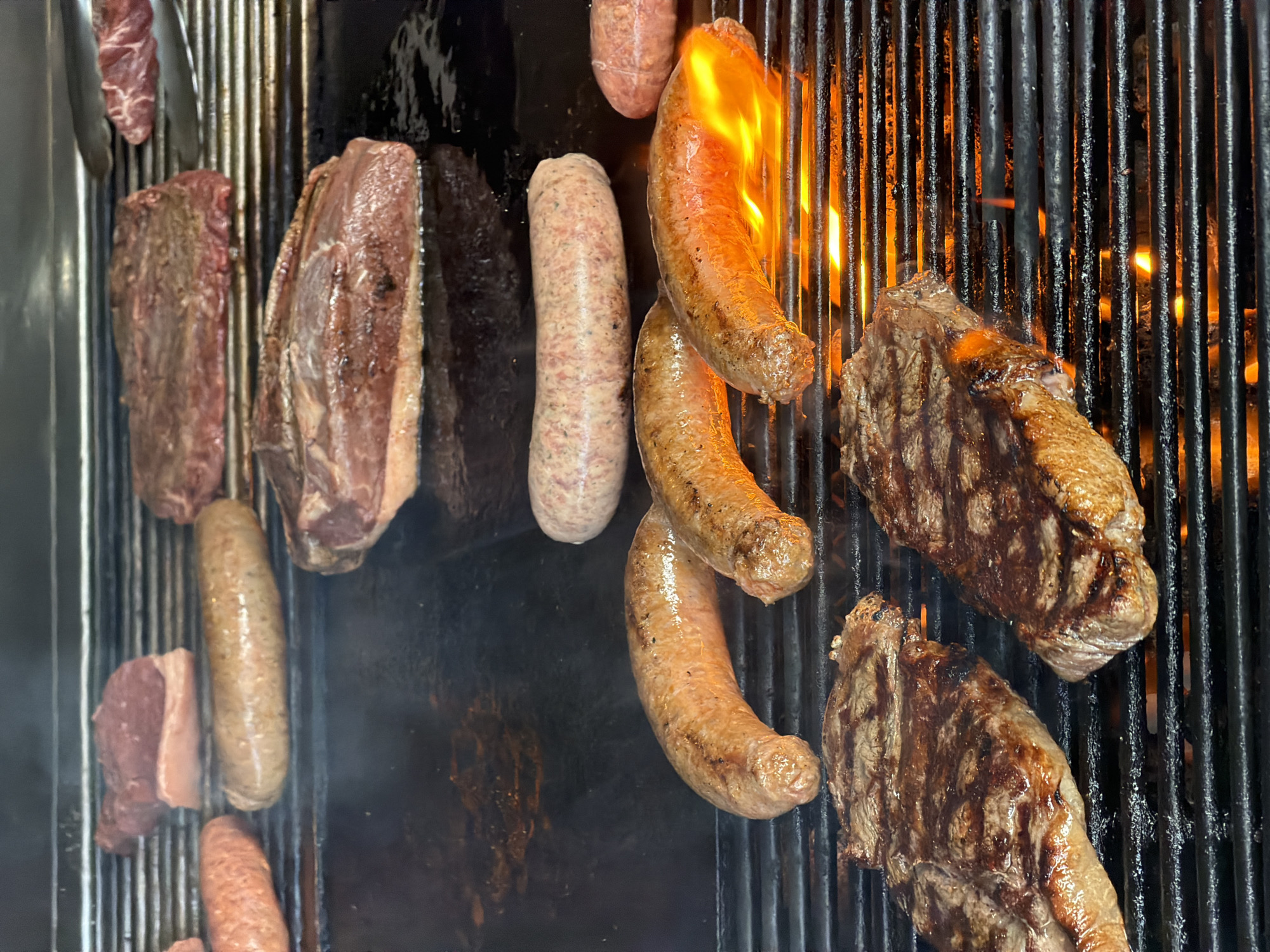 The image shows a grill with various meats and sausages being cooked. There are several pieces of steak, some with visible grill marks, and an assortment of sausages, including what appears to be a lighter-colored type and some that are browning and even flaming. The grill is made of black metal bars. The image was taken in Kerobokan, Indonesia.