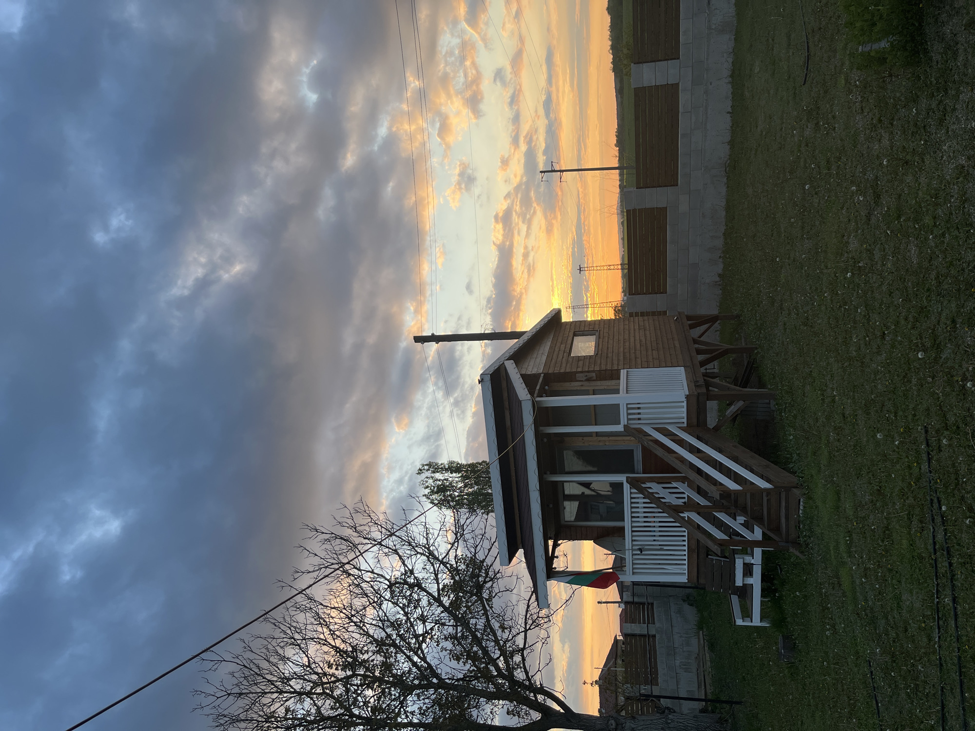 The image shows a one-story wooden house with a white railing and stairs, set against a dramatic sky with dark clouds on one side and a vibrant sunset on the other. A bare tree is in the foreground, partially obscuring the house. Power lines stretch across the sky. A Bulgarian flag hangs from the house's porch. The house is situated in a grassy area, with a fence visible in the background. Based on the request, the location is identified as Pchelnik, Bulgaria. There are no people visible in the image.