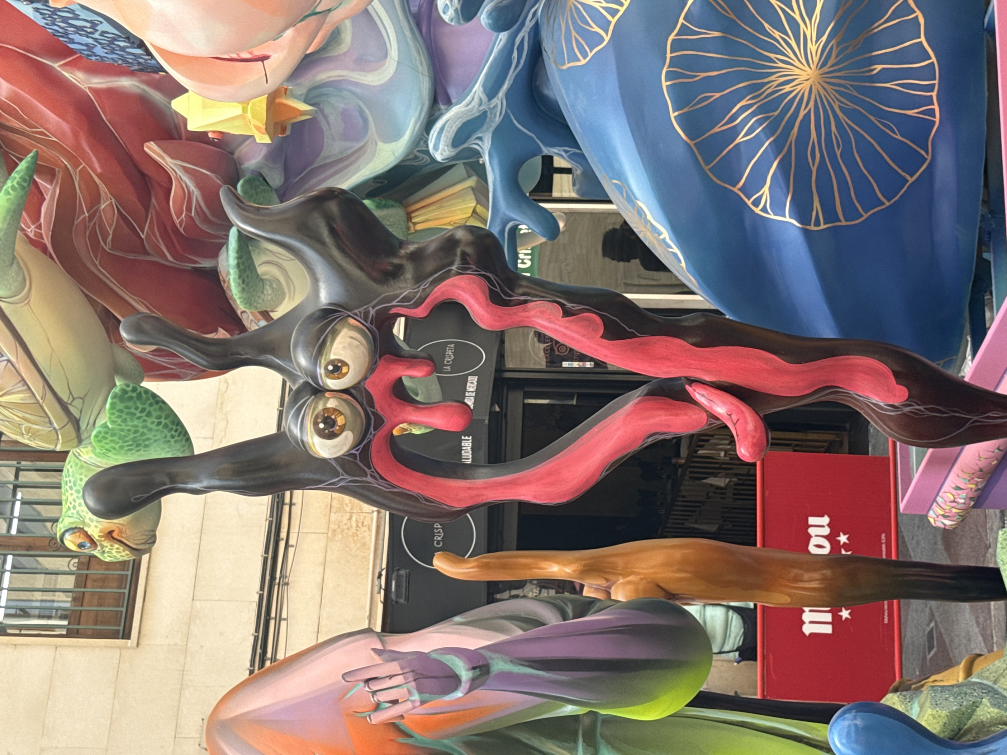 A close-up, eye-level shot captures a vibrant, surreal sculpture with exaggerated features. A grotesque figure with wide, bulging eyes, dark, slicked-back hair, and a gaping mouth with long, red, fleshy tongues dominates the foreground. The sculpture's body is adorned with thin, vein-like red lines. Behind this central figure, a bright green cartoonish turtle with a patterned shell is visible, its head turned to the left. Further in the background, a large, blue, rounded object with intricate gold line art resembling a stylized flower or sunburst is prominent. To the left, a tall, slender, brown, vaguely hand-like sculpture stands upright. A red banner with white lettering and two small stars is partially visible at the bottom left, with the text "mou" clearly legible. Other abstract, colorful sculptural elements, including a pale pink form with a purple hand and an orange form, are also present, creating a chaotic and artistic composition. The background shows a building facade with windows and some commercial signage, suggesting an outdoor urban setting.