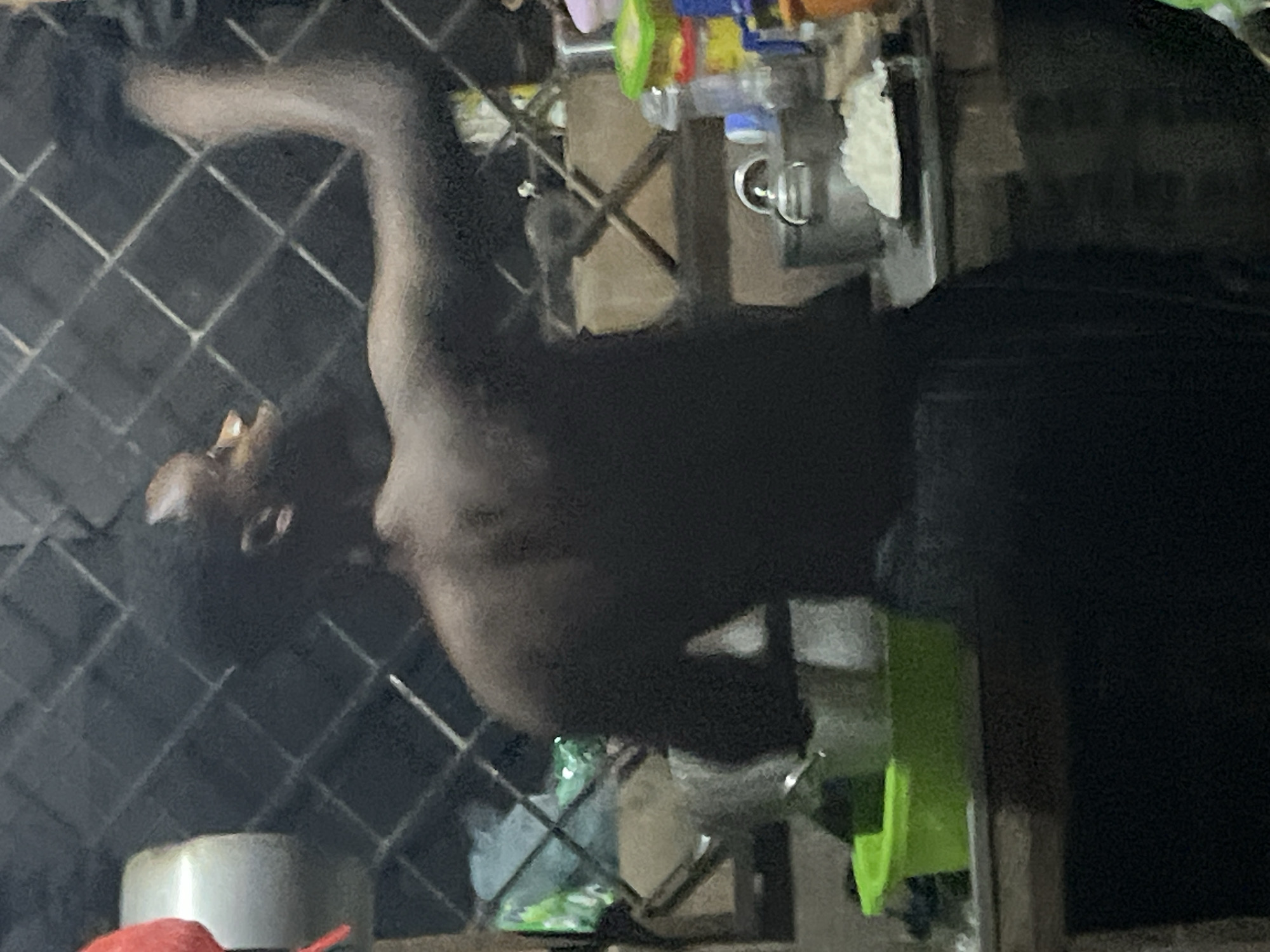 The image shows a person's torso and arm, with their head mostly out of frame, positioned behind a diamond-shaped mesh grate. The person appears to be shirtless, with dark hair. They are looking downwards and to the left. The background is dimly lit and cluttered, with some shelves visible. On the shelves, there are various colorful objects, possibly toys or kitchen utensils, and some white fabric. A metal handle is also visible on the right side of the frame. The overall scene appears to be indoors, possibly a kitchen or storage area. The lighting suggests it might be evening or a poorly lit indoor space. There is no visible text in the image.