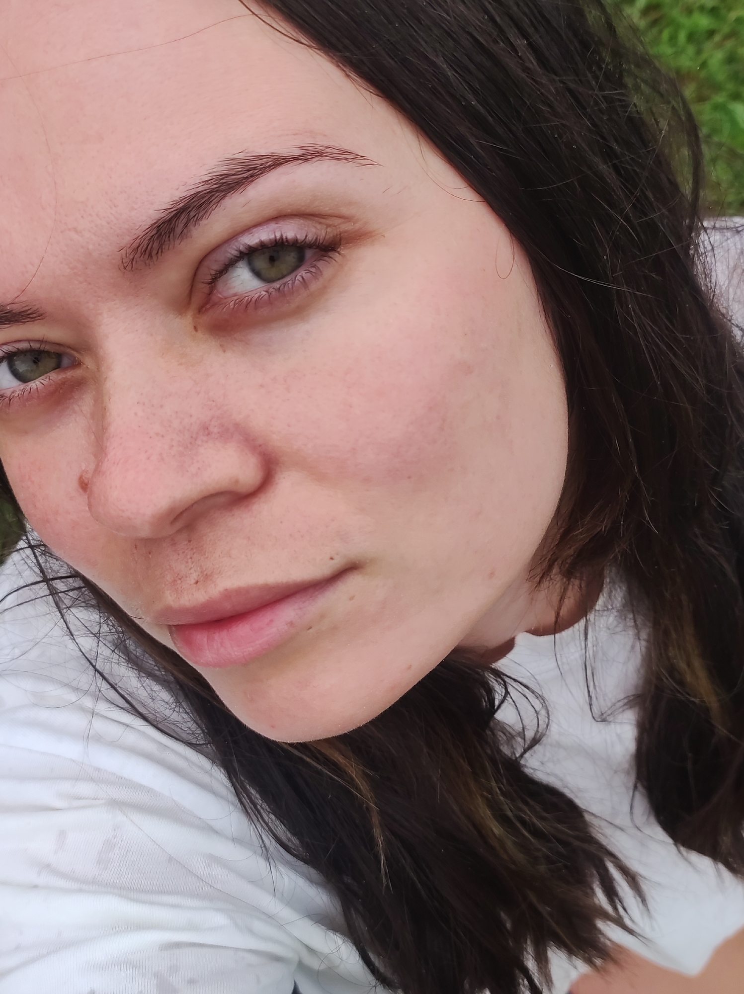 A close-up, high-angle shot shows a fair-skinned person with dark brown hair and green eyes. The person is wearing a white shirt. The background appears to be an outdoor setting with grass visible in the periphery. The person is not smiling, and the expression is neutral. The person has freckles on their nose and cheeks, and visible eyebrow hair. There is no indication in the image of the city of Фоки, Russia.