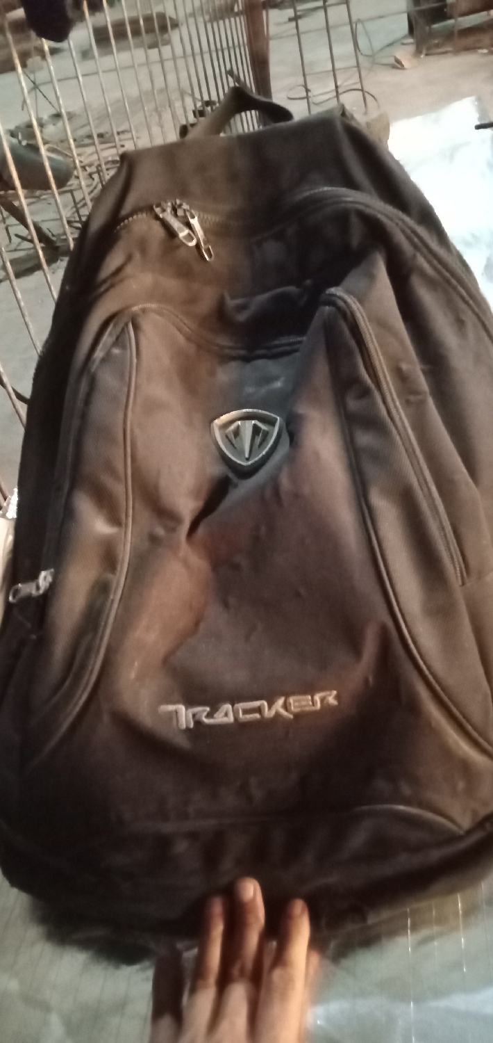 The primary subject of the image is a dark-colored backpack, likely black or dark grey, positioned centrally. The backpack is made of fabric and displays signs of regular use, with some dust or faint marks on its surface. It features multiple compartments, indicated by visible zippers; a main compartment on top has two metallic zipper pulls.
On the upper front of the backpack, there is a metallic emblem shaped like a shield, containing a stylized design resembling the letter "T." Below this emblem, the brand name "TRACKER" is clearly visible, written in a light-colored, embossed or embroidered uppercase font across the lower front pocket.
At the bottom of the frame, a human hand with visible fingers is present, resting near or against the base of the backpack. There is no discernible activity other than the hand's presence in the shot.
The setting appears to be an indoor or semi-enclosed space, possibly a functional or storage area. The background, which is slightly out of focus, shows metal bars or grating, suggesting structures such as shelving, a cage, or a security barrier. The lighting conditions are somewhat dim, indicating either artificial illumination or low natural light. The backpack rests on a light-colored, smooth surface, which could be a floor or a workbench. The general impression is of a casual, non-formal environment. The location context is Sumput, Indonesia.