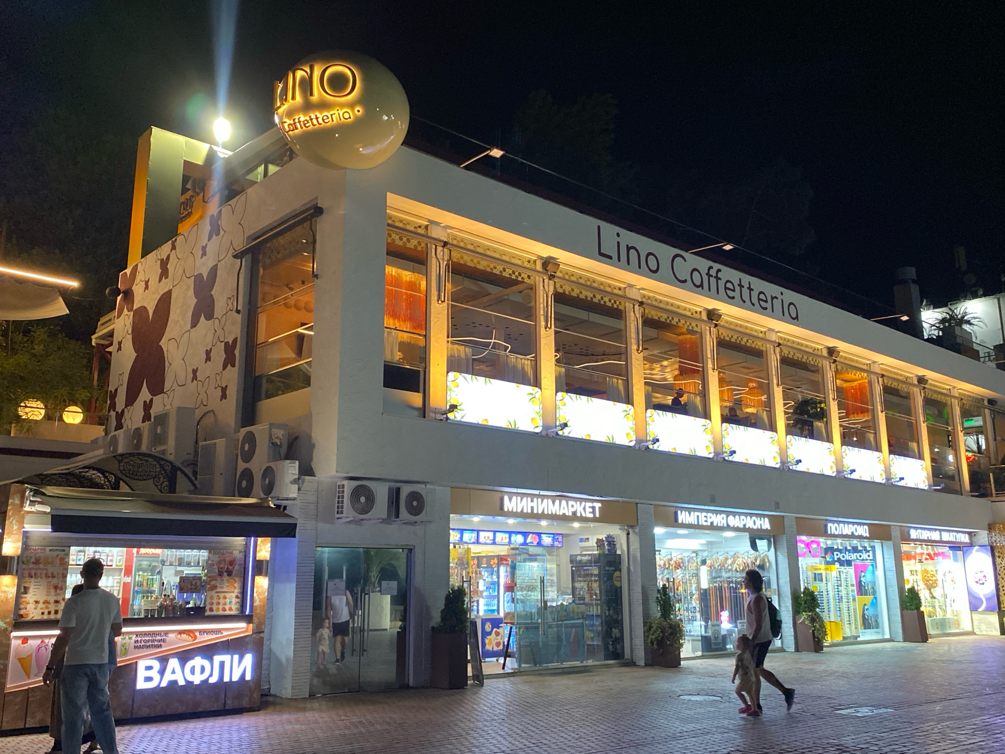 The image captures a nighttime scene in Sochi, Russia, specifically on a pedestrian walkway in front of a commercial building. The building features several storefronts and a second-floor establishment with large windows illuminated from within.
On the ground level, to the left, is a food stall with bright lighting displaying various beverages and waffle items. A man wearing a white t-shirt and blue jeans stands with his back to the camera, looking towards the food stall.
Further to the right, there are multiple shops. One is labeled "МИНИМАРКЕТ" (Minimarket), another "ИМПЕРИЯ ФАРАОНА" (Empire of the Pharaoh), and one "ПОЛАРОИД" (Polaroid). A woman wearing a white tank top and dark shorts, carrying a backpack, is walking to the right with a young child. The child is wearing a light-colored dress.
The building's facade is decorated with floral patterns in reddish-brown and white. A prominent round sign with illuminated text "LINO Caffetteria" is visible above the second floor. The second floor itself has large windows with yellow lighting and decorative elements.
The walkway is paved with patterned tiles. Trees and potted plants are placed along the storefronts, and air conditioning units are mounted on the building's exterior. The overall impression is a lively commercial area at night.