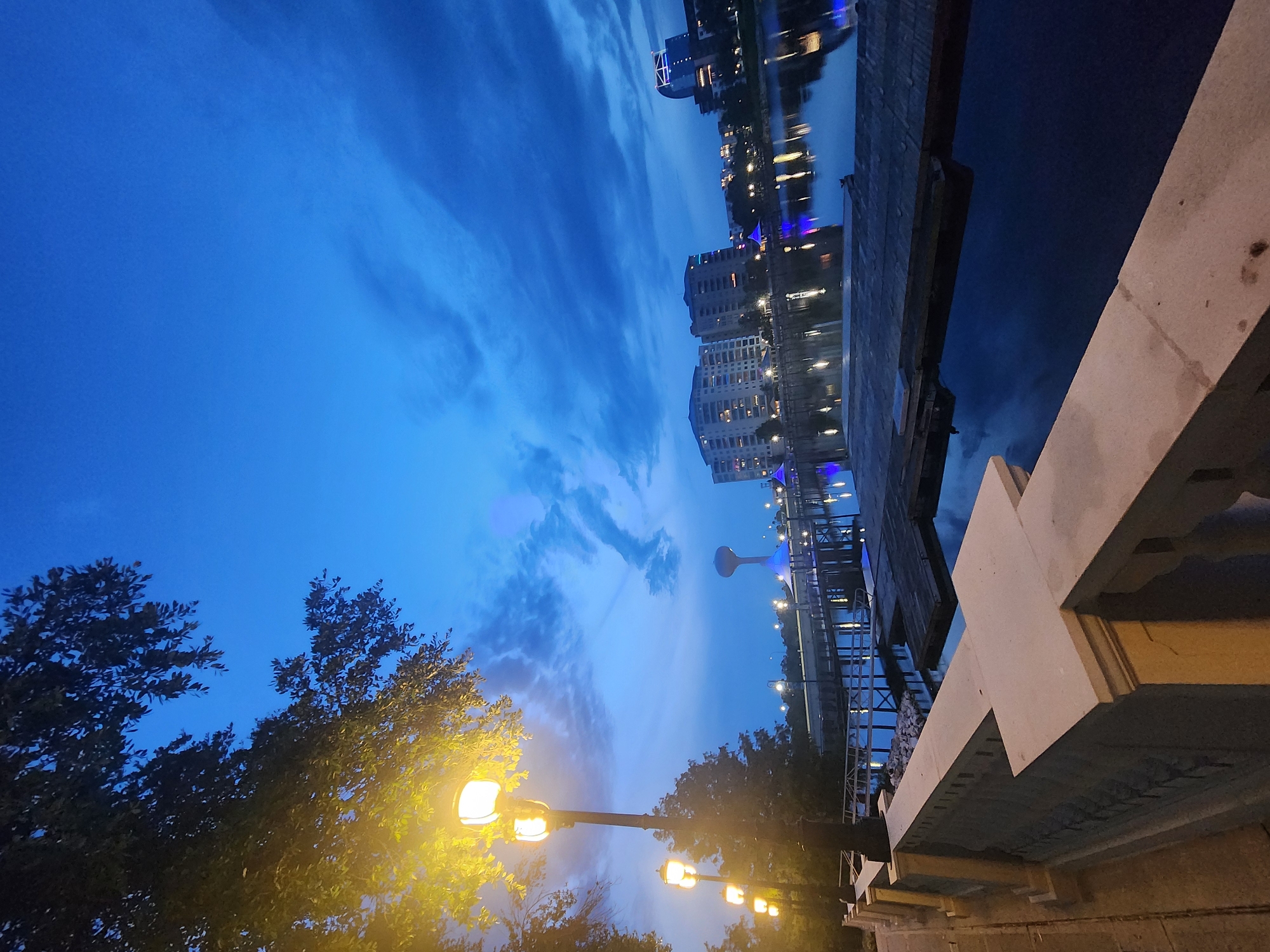 This is a vertical, eye-level, medium shot of the Altamonte Springs lakefront at dusk. The sky is a deep, vibrant blue with scattered, wispy clouds. In the foreground, a beige, stone railing lines the walkway, with a tree branch illuminated by a warm, yellow lamp post extending from the left side of the frame. The walkway leads to a body of water, likely part of the Crane's Roost Lake, reflecting the lights of the city.
Across the water, a long pier with several lamp posts stretches into the distance. Beyond the pier, a water tower with a blue-lit base stands tall. Further in the background, several modern buildings with illuminated windows are visible, casting reflections on the water. The overall atmosphere is peaceful and serene, showcasing the city's urban landscape at twilight.