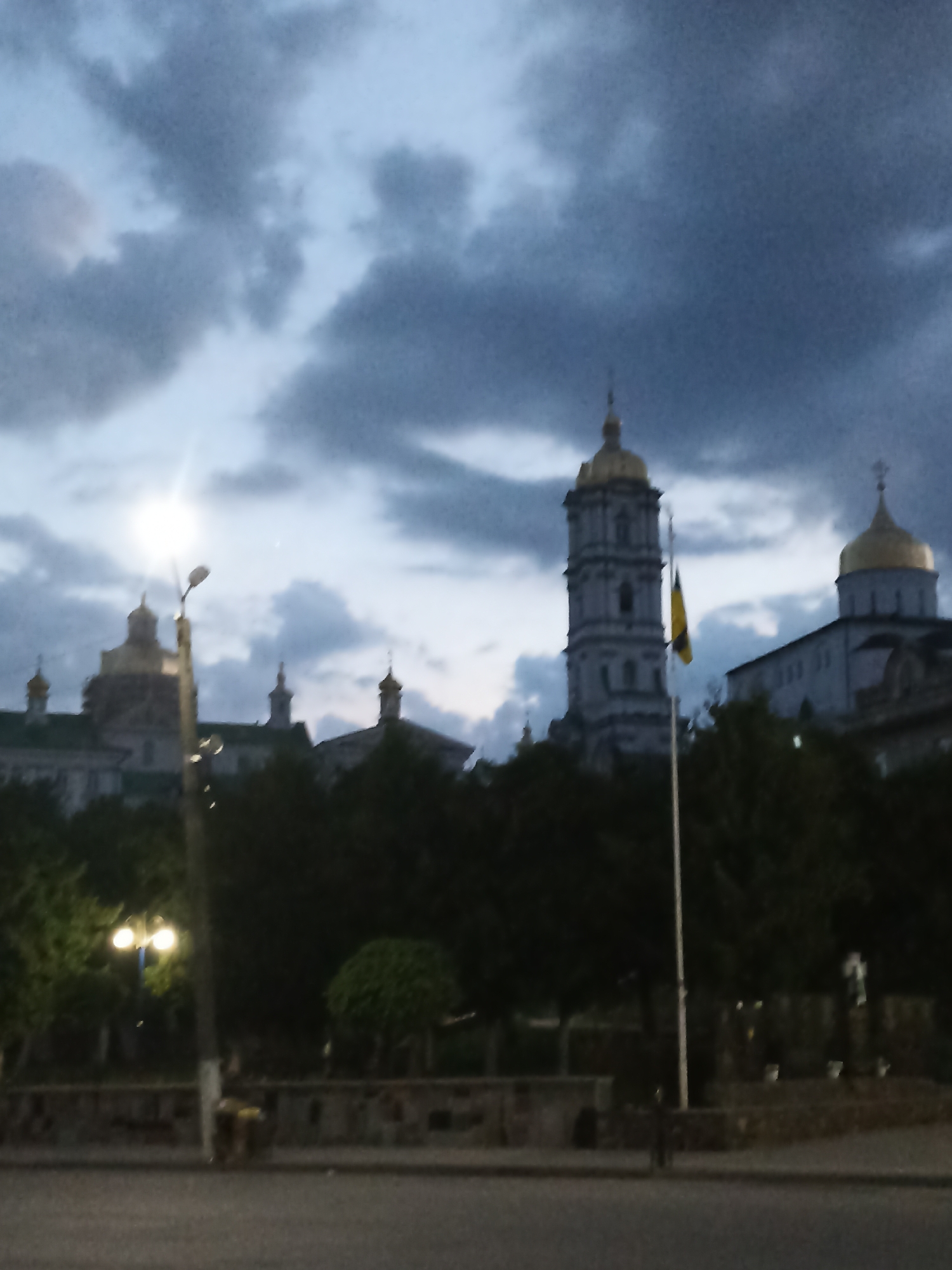 The image depicts a somewhat blurred view of the Pochaiv Lavra in Ukraine, captured during what appears to be dusk or early evening. The sky is overcast with dramatic, dark clouds, suggesting a moody or possibly stormy atmosphere.
Prominent in the background are the distinctive golden domes of the monastery's churches, including a tall, multi-tiered bell tower. A Ukrainian flag is visible, flying on a flagpole in front of the bell tower, adding a patriotic element to the scene. In the middle ground, a line of trees partially obscures the lower portions of the monastery complex.
In the foreground, a street light with two bright bulbs is illuminated, casting a glow and a lens flare that partially washes out the detail of the lamp post. The light suggests that evening has fallen or is approaching. The ground in the immediate foreground appears to be a paved surface, possibly a road or plaza. The overall impression is one of a solemn and historic location at a transitional time of day, with a slightly somber and dramatic sky. No people are clearly discernible in the image, and no text is visible.