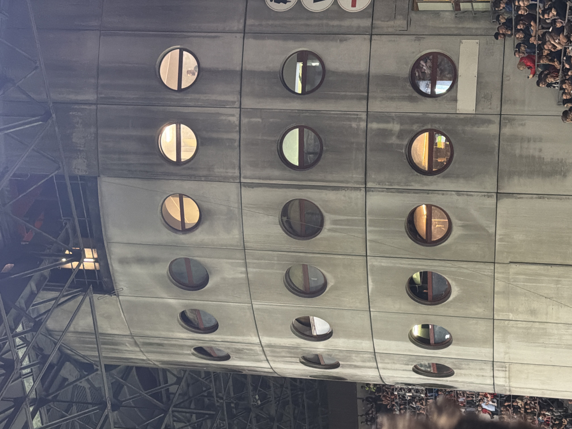 The image shows a section of the Stadio San Siro in Milan, Italy. The stadium's exterior is characterized by a gray, modular concrete structure with a grid of circular windows. A scaffolding structure is visible on the left side of the image. A crowd of people is visible on the right side of the image, partially obscured.