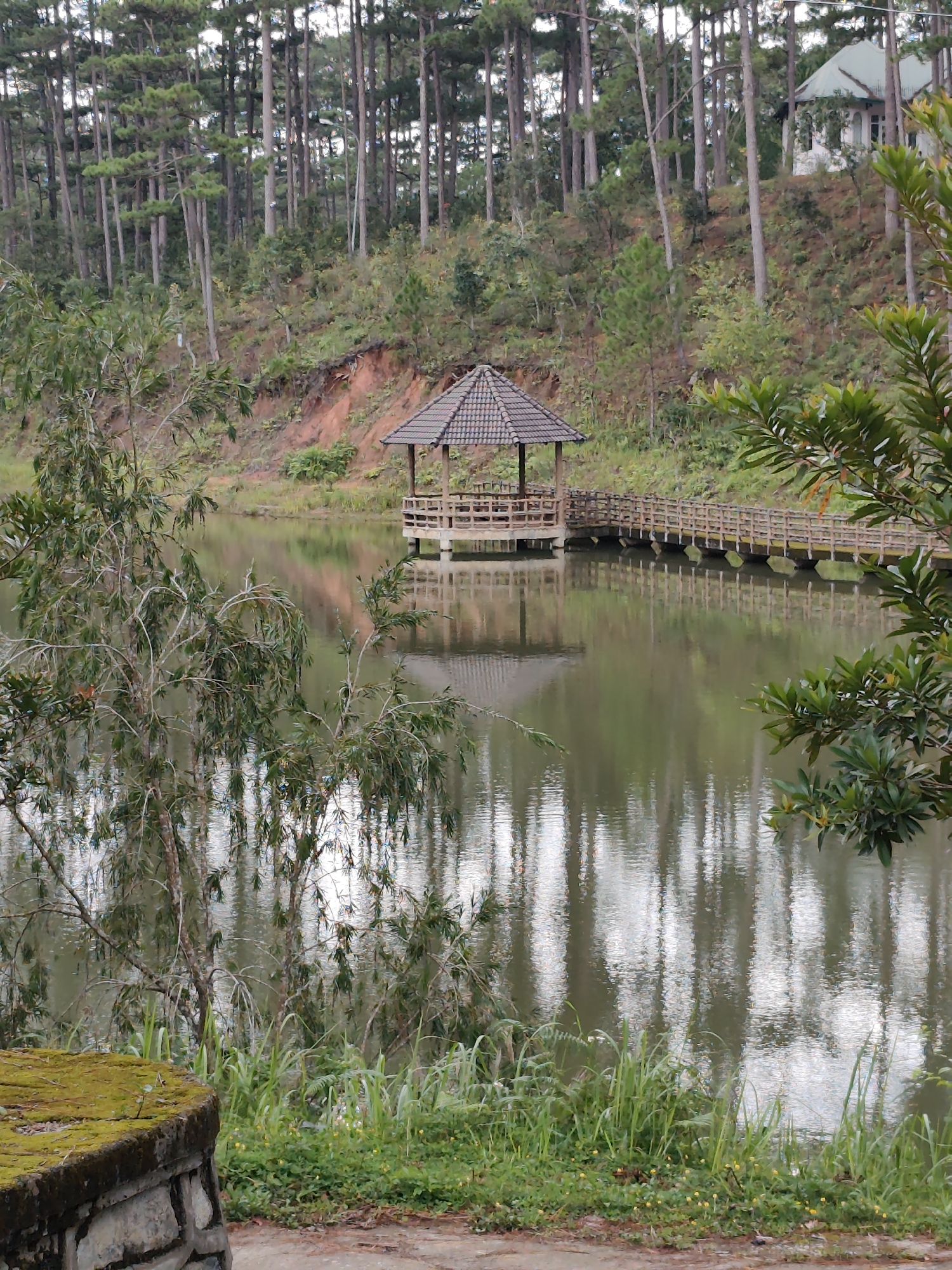 A picturesque lake scene features a wooden gazebo and a bridge. The gazebo is situated on the left side of the lake, with a sloped roof and wooden posts supporting it. A wooden bridge with railings extends from the gazebo across the water, with its reflection shimmering on the lake's surface. The lake is surrounded by lush green trees, some of which are reflected in the water, creating a serene and tranquil atmosphere. In the background, a white building with a green roof is visible, nestled among the trees. The image has a soft focus, giving it a dreamy and painterly quality.