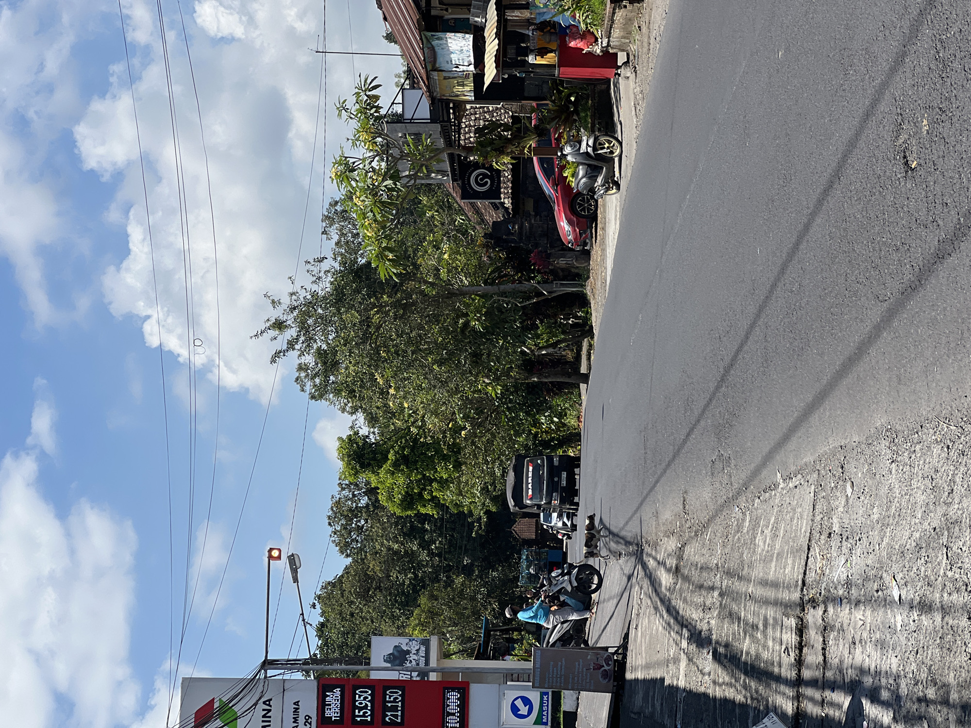 The image depicts a street scene in Payangan, Indonesia, on a sunny day with scattered clouds. On the left, a Pertamina gas station sign is visible, displaying fuel prices including "BELUM TERSEDIA" and "15.950" and "21.150". A person on a white scooter is stopped near the gas station, while a dog walks across the road.
Further down the road, a black truck with "BARES" written on its side is driving away. To the right of the road, there are several buildings with rustic roofs and storefronts, including what appears to be a warung (small shop) with food items displayed. A red car is parked in front of one of the buildings, and a dark scooter is parked in front of another. Trees and foliage line the sides of the road, contributing to a tropical environment. Power lines are strung across the sky above the scene. The road surface shows some wear and tear, with a pothole visible in the foreground.