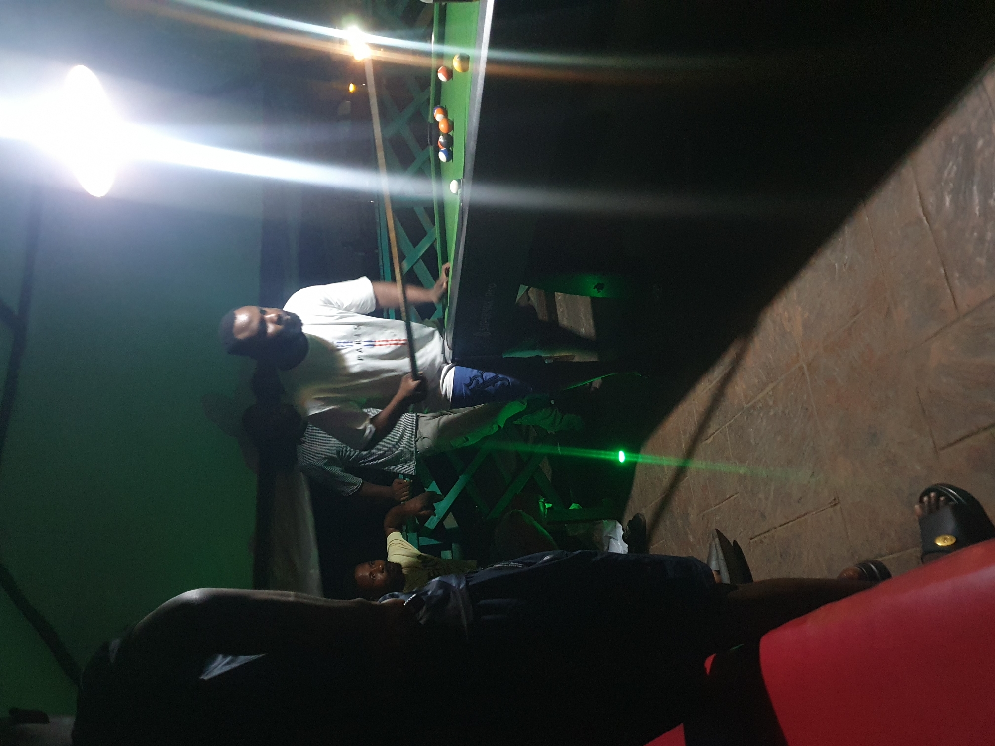 In Lokoja, Nigeria, at night, a group of men are gathered around a pool table in an outdoor setting, possibly a bar or recreational area. The scene is illuminated by a bright overhead light, casting strong shadows. On the pool table, several balls are visible, including a cue ball, and various colored billiard balls. One man, wearing a white t-shirt with "PARIS" printed on it, is holding a pool cue, appearing to be in the process of playing. Another man in a yellow t-shirt is leaning on a green wooden structure near the table. The ground appears to be paved with stone tiles. A red seating area is partially visible in the foreground. The overall ambiance suggests a casual evening of recreation.