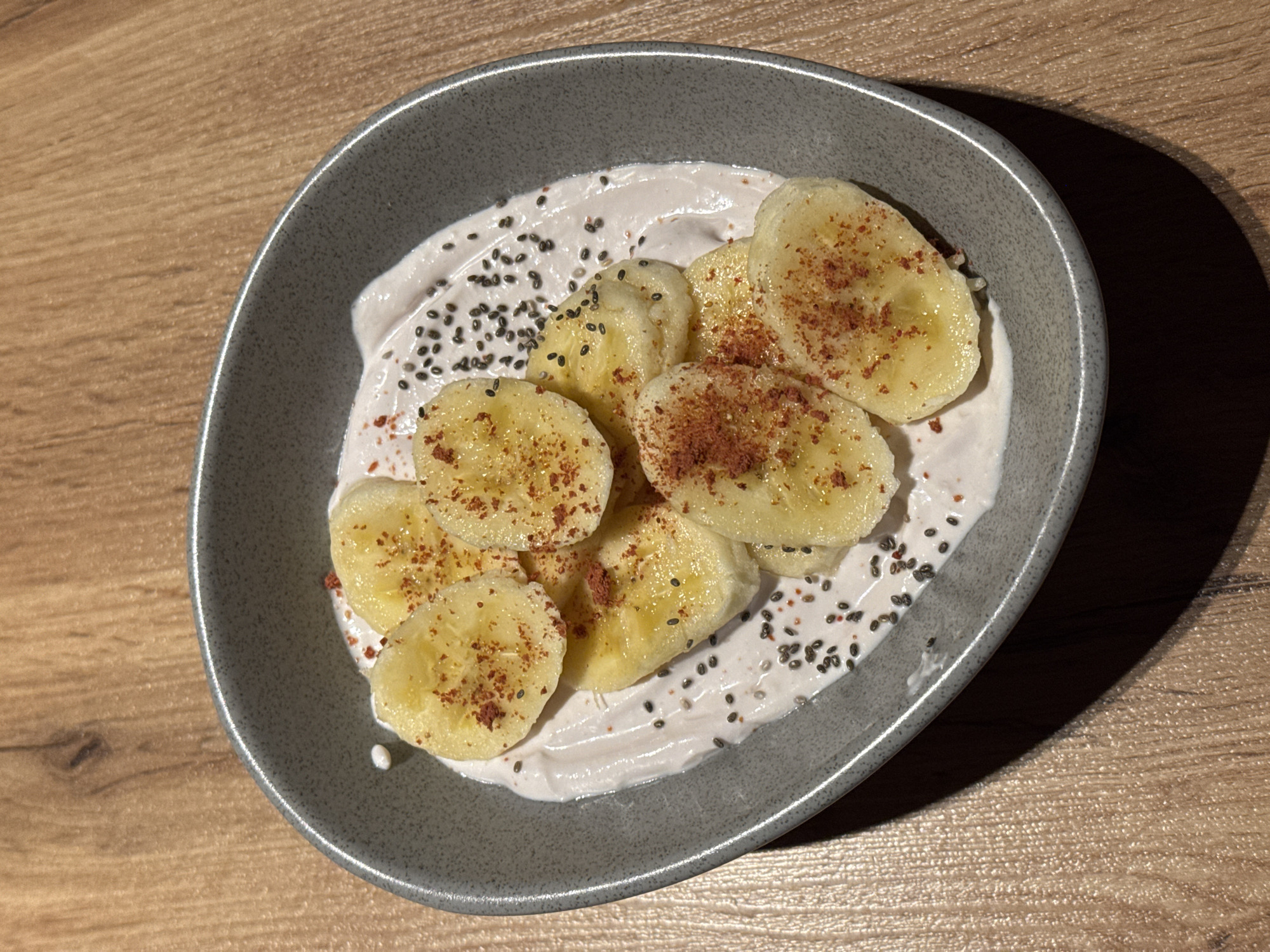 A grey, organically shaped ceramic bowl containing a food item is positioned on a light brown wooden surface with visible grain. The bowl holds a creamy, pale pinkish-white base, likely yogurt, topped with multiple slices of ripe yellow banana. Scattered over the creamy base and between the banana slices are small, dark chia seeds. A fine, reddish-brown powder is sprinkled unevenly across the banana slices. A distinct shadow from the bowl is cast on the wooden table towards the lower right, indicating an overhead light source from the upper left. No people or other objects are visible.