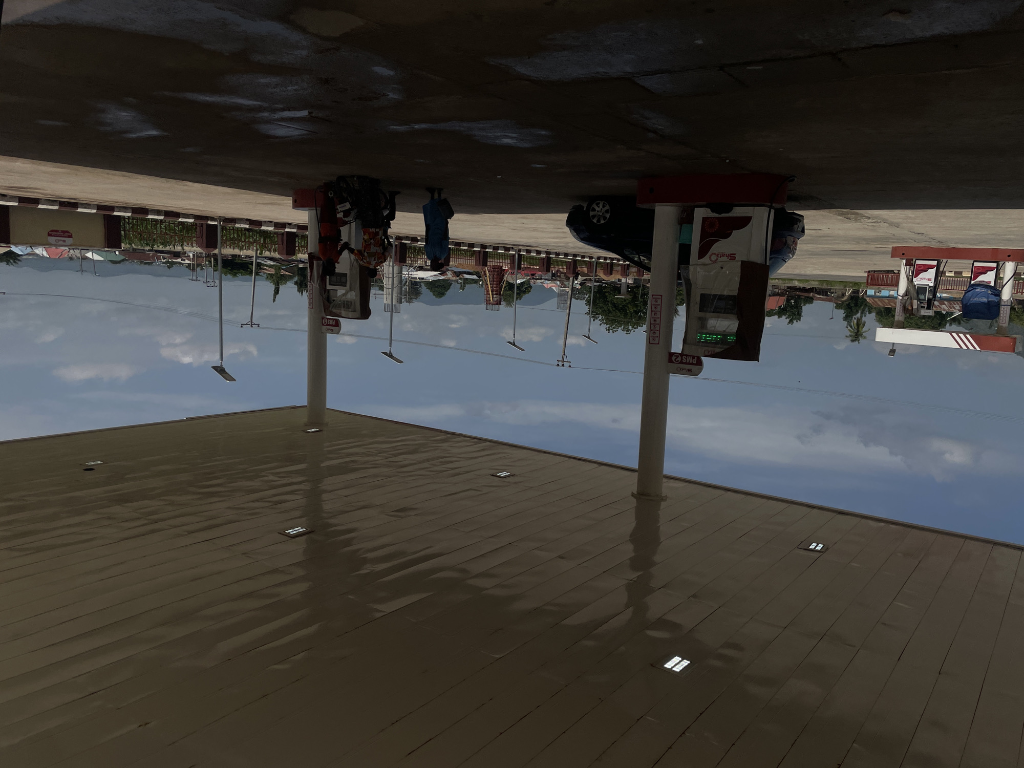 This is an outdoor scene at a Shell gas station in Bassa, Nigeria. The image is oriented sideways. In the foreground, there is a large, flat, light brown roof with several square lights embedded in it. The roof reflects the sky. In the middle ground, there are two Shell-branded gas pumps. One pump has a blue bag on it, and a black car is parked next to it. Two people, one wearing a blue dress and carrying a bag, and another person in an orange and patterned shirt, are standing near the pumps. The other gas pump appears to be unused. In the background, there are trees, buildings with red and white striped accents, and mountains in the distance under a cloudy blue sky. The overall impression is a normal day at a gas station. There is no visible text in the image.