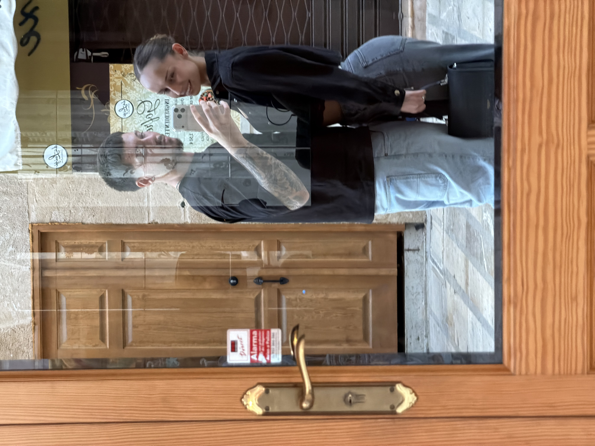 A couple is reflected in the glass door of a business in Alcúdia, Spain. The male, wearing a black t-shirt and light blue jeans, is holding up a white smartphone. A woman in a black jacket and light blue jeans stands beside him. The reflection shows the business's interior, including a wooden door, and a security system sticker. The exterior wooden door frame and handle are in the foreground. The reflection indicates they are taking a photo of the business or perhaps a selfie with the business in the background.