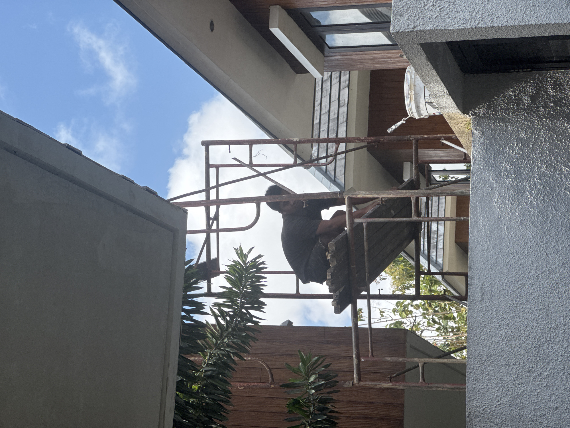 A man wearing a black t-shirt and shorts is crouching on a wooden plank as part of a metal scaffolding structure. He appears to be working on the exterior of a building. To his right, a white bucket with a paint roller is placed on a ledge. The building features brown wooden paneling and a gray, textured wall in the foreground. The sky above is blue with scattered white clouds. This scene is situated outdoors in Kuta Selatan, Indonesia.