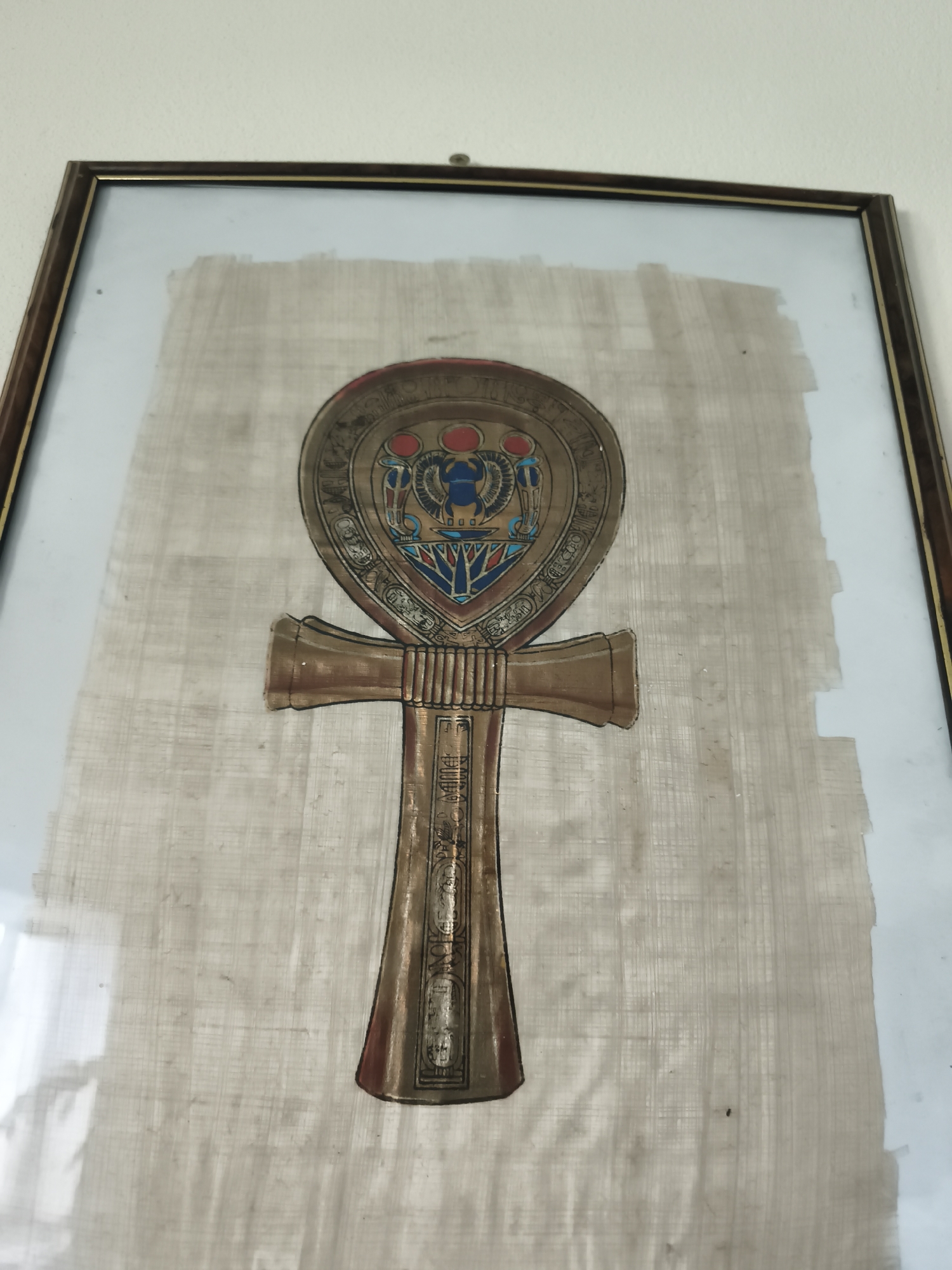 A framed Egyptian Ankh symbol painted on papyrus hangs on a wall in a room. The Ankh is a cross with a loop at the top, and it is decorated with hieroglyphs and depictions of figures. The papyrus is off-white and has a textured appearance. The frame is dark brown with a thin gold trim. The wall is painted white. The image was taken indoors, likely in a residential setting. There are no people or animals visible in the image. The lighting is natural, coming from an unseen window, casting reflections on the glass of the frame.